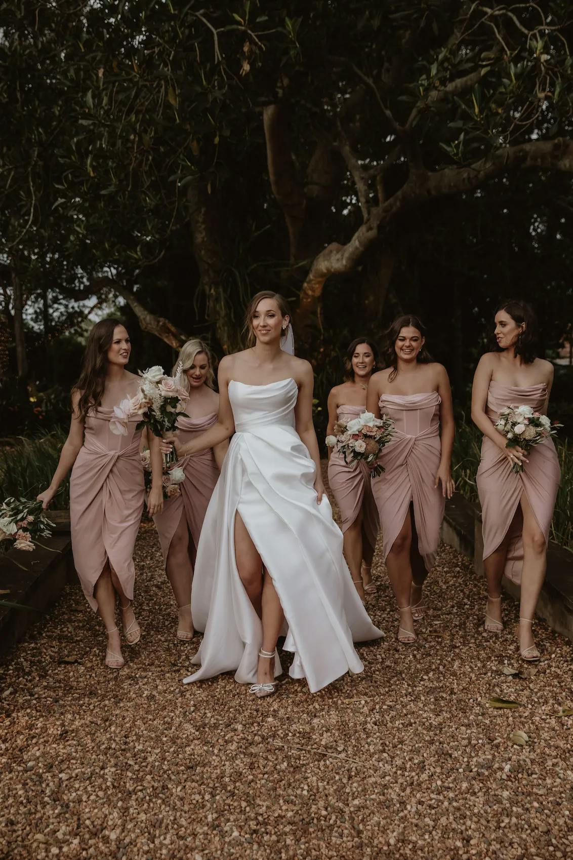A bride in a white wedding dress walks down a gravel path, accompanied by five bridesmaids wearing matching soft pink dresses. Each bridesmaid holds a bouquet of flowers. They are all smiling and appear to be having a joyful moment together. Trees and foliage are in the background.