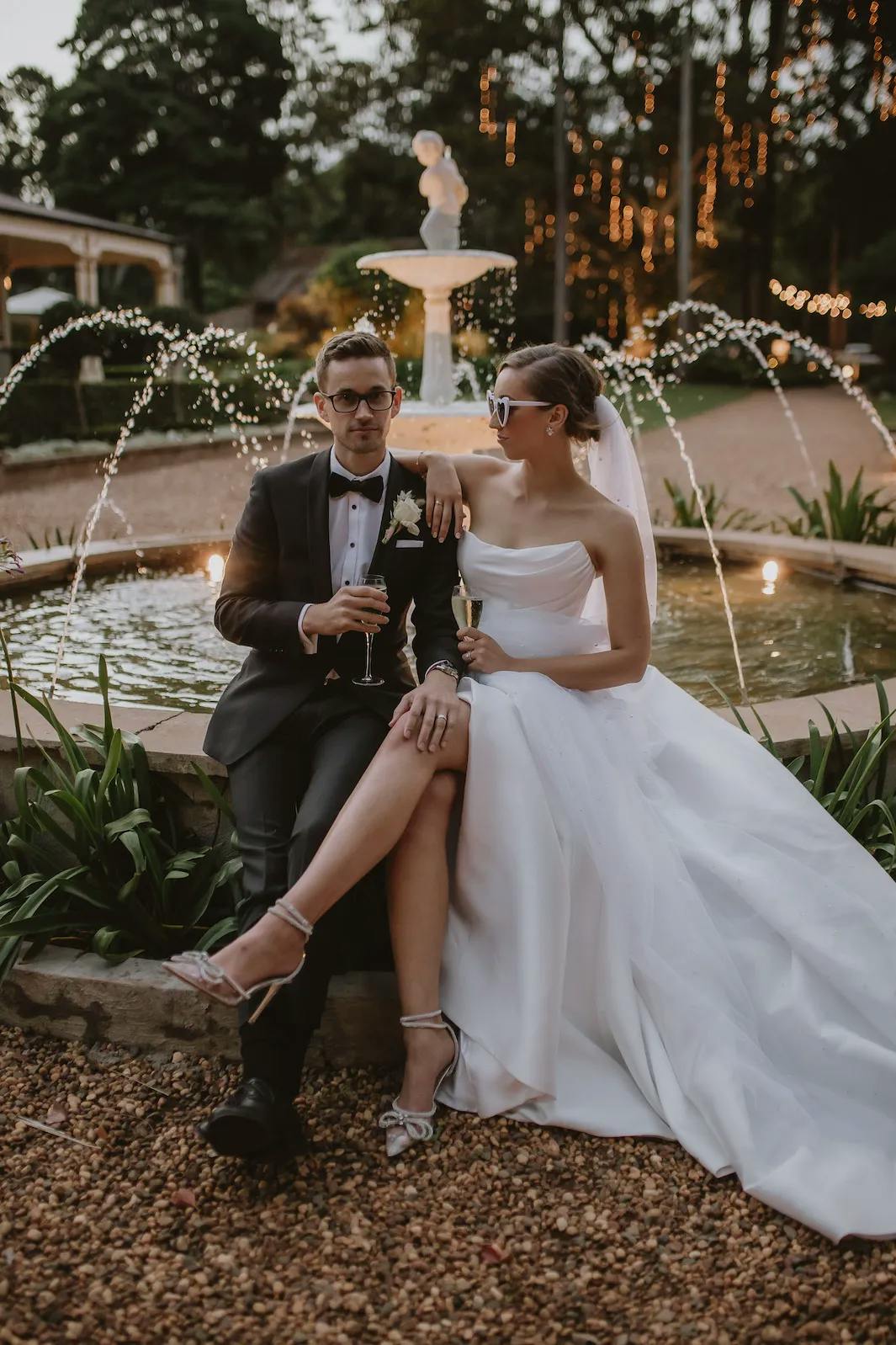 A bride and groom sit together on the edge of a stone fountain in an outdoor setting. The groom wears a black tuxedo, and the bride is in a white strapless wedding gown with a veil. They both hold champagne glasses, and the bride is wearing sunglasses. Fairy lights are visible in the background.