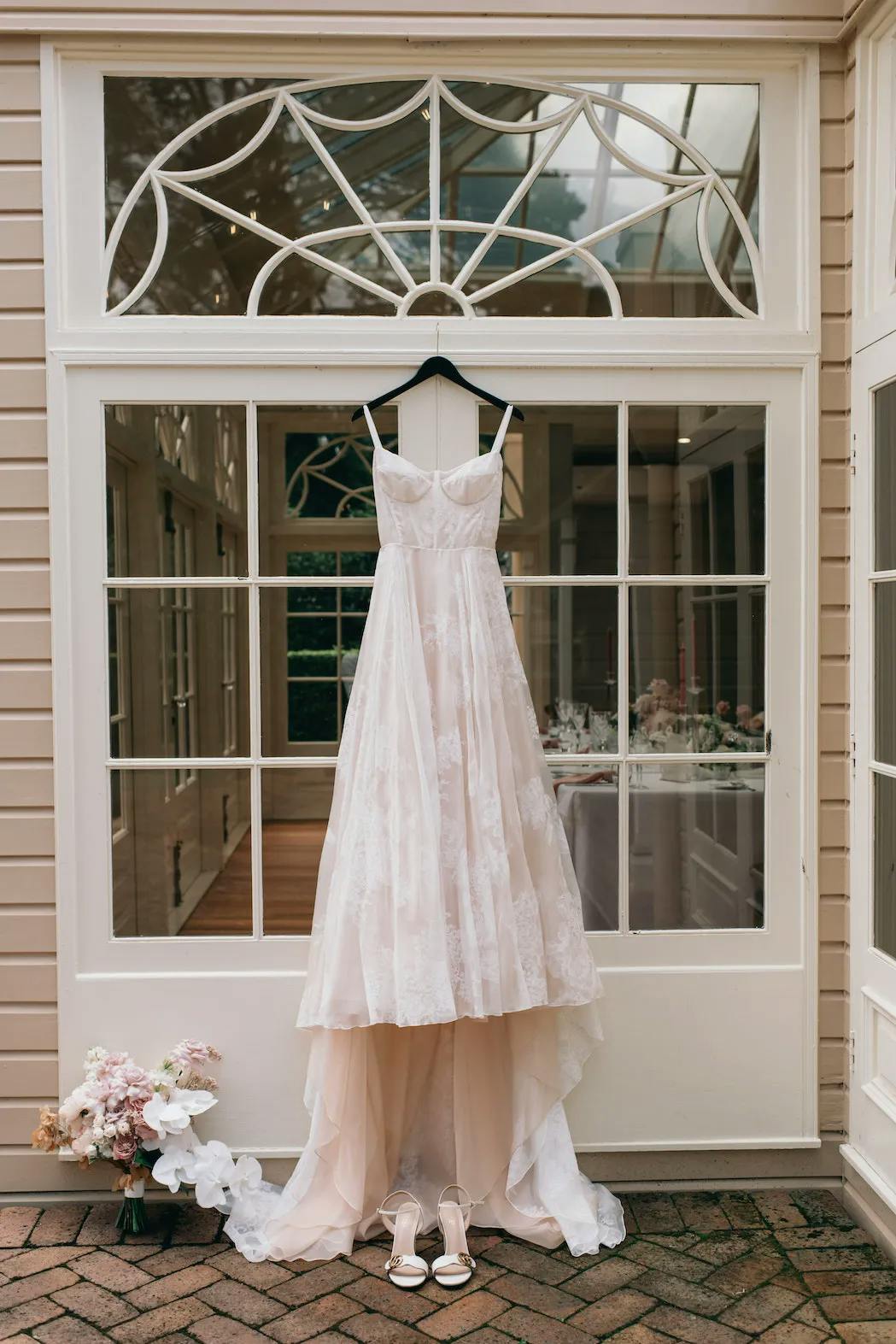 A white wedding dress is hanging from a window by thin straps, displayed in front of a glass door with decorative framing. Below the dress, a pair of white high-heeled shoes are placed on the ground next to a bouquet of pink and white flowers.