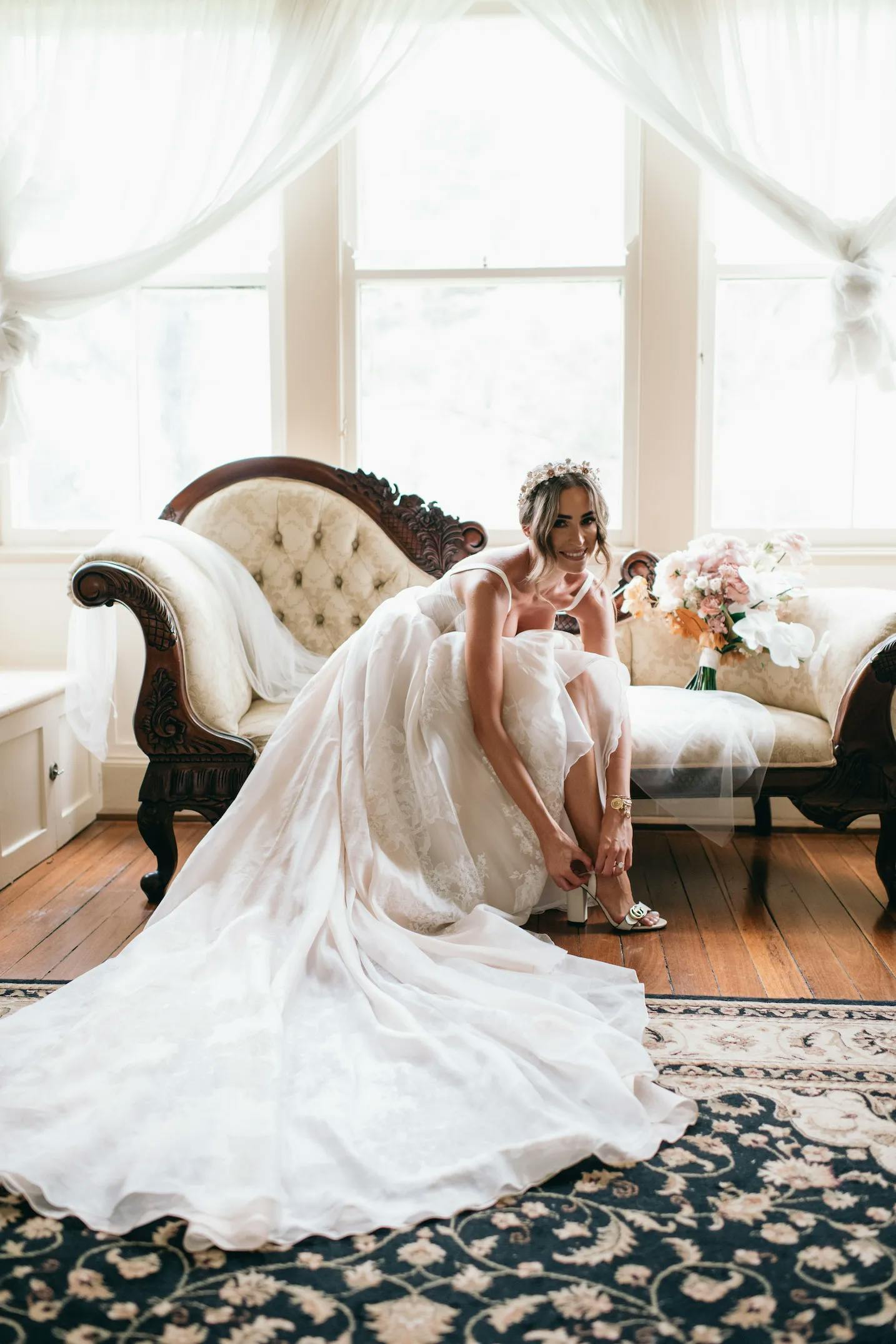 A bride in a white wedding gown sits on an antique sofa near a window with sheer curtains. She adjusts her white high-heel sandal while smiling. A bouquet of flowers is placed on the sofa beside her, and an ornate rug covers the wooden floor.