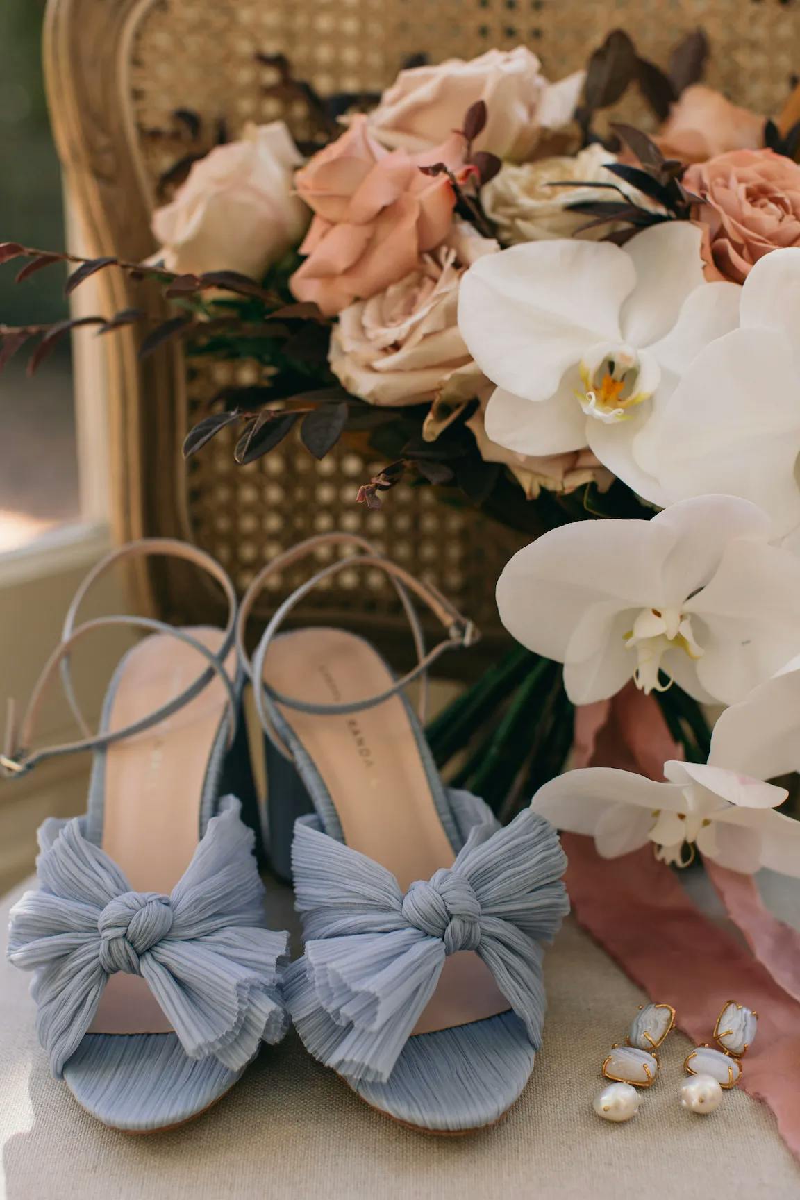 A pair of elegant blue high-heeled shoes with bow accents, placed in front of a lush bouquet of flowers featuring white orchids and peach-colored roses. The scene is completed with a pair of dangling gold and pearl earrings.