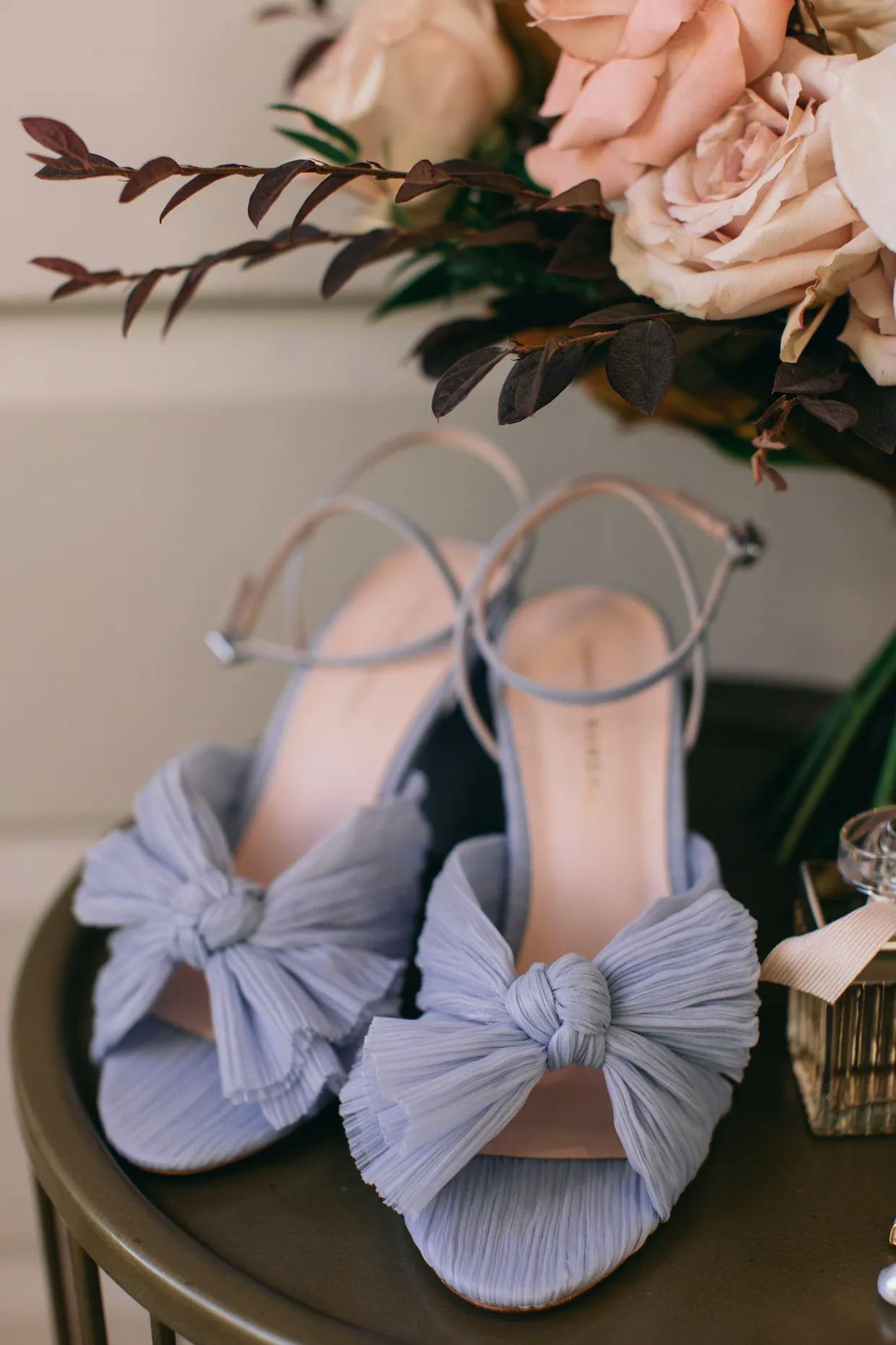 A close-up image of light blue high-heeled sandals with large fabric bows on the front. The sandals are displayed on a round table next to a bouquet of pastel roses and greenery and a small bottle of perfume. The background is softly blurred.