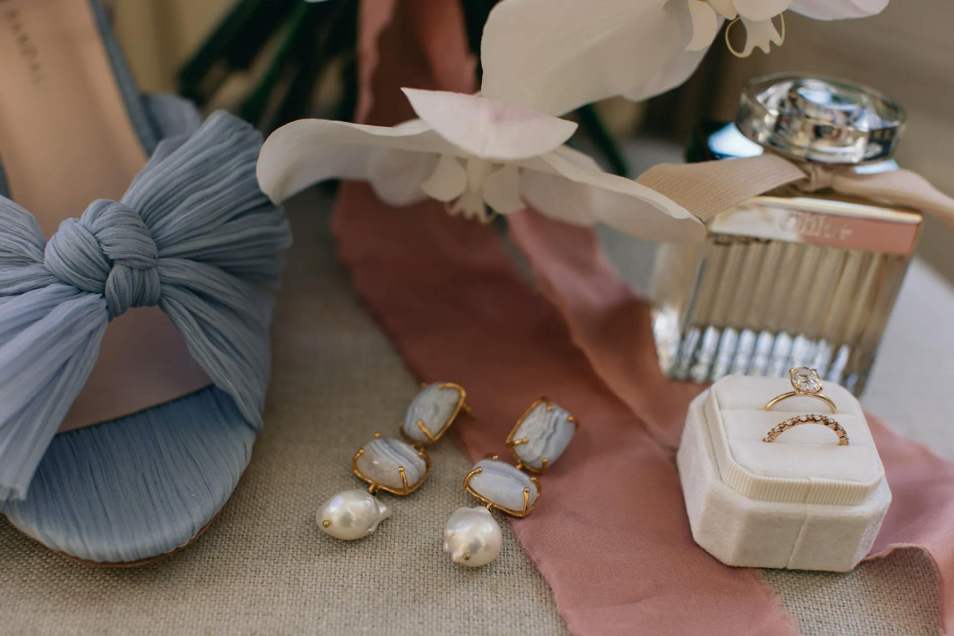 A close-up of wedding accessories includes a blue shoe with a bow, a pair of gemstone and pearl earrings, a white ring box containing two rings, and a perfume bottle placed on a beige surface with a pink ribbon. White flowers are partially visible in the background.