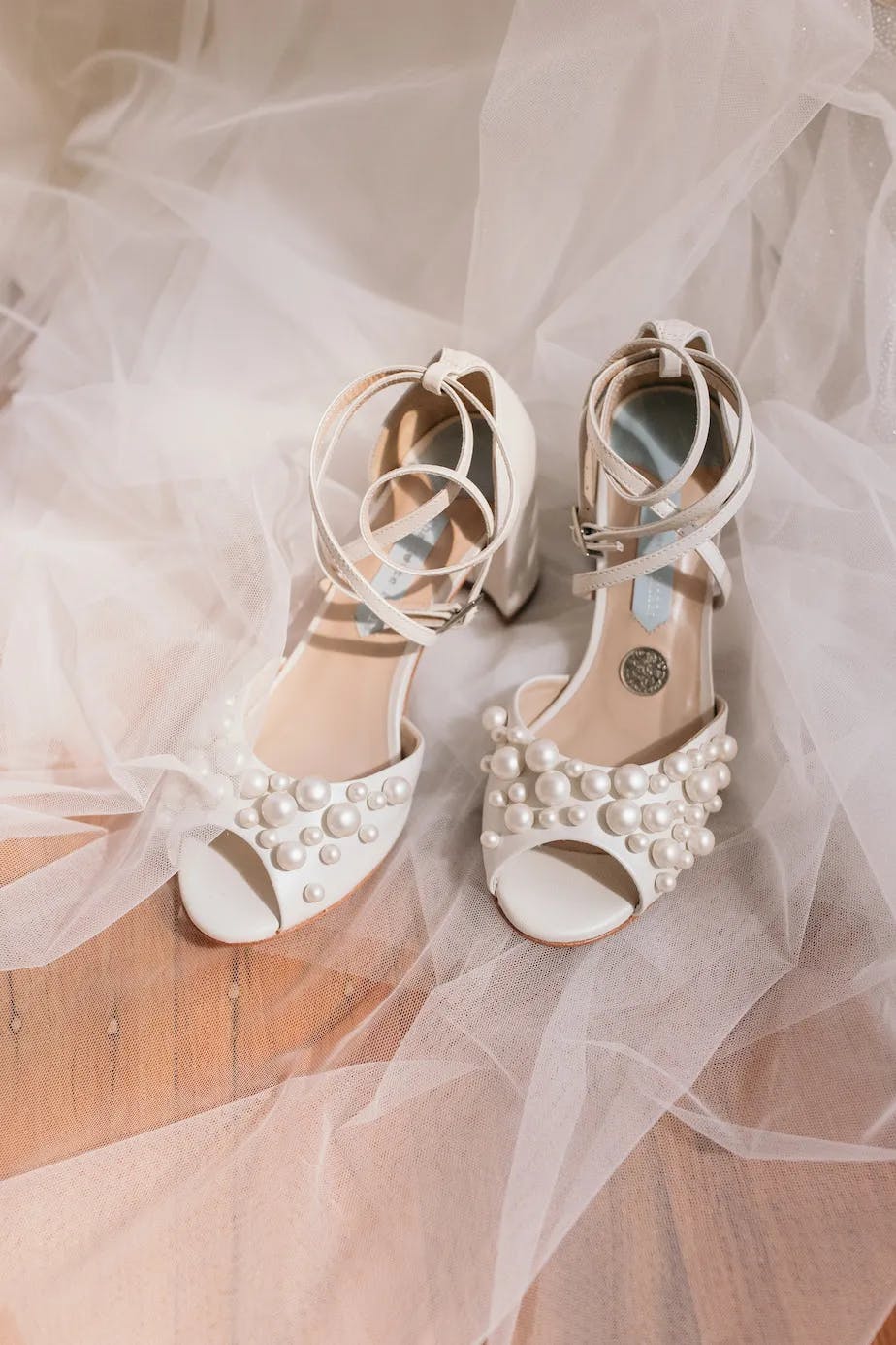 A pair of elegant white high-heeled shoes adorned with pearls and ankle straps placed on a wooden floor, with sheer tulle fabric softly draped around them, creating a delicate and dreamy atmosphere.