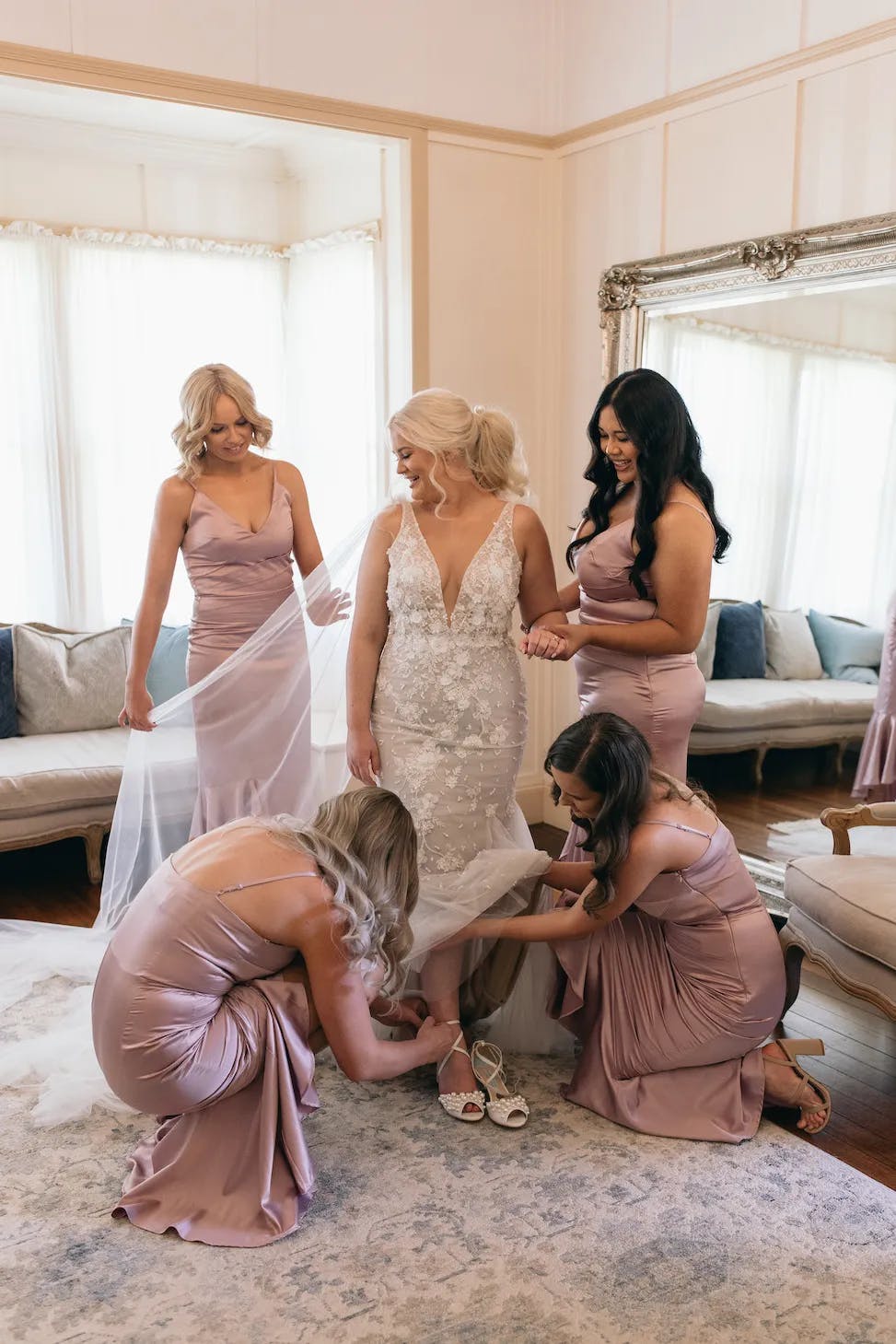 A bride in a lace wedding dress stands in the center of a room, surrounded by four bridesmaids in matching light purple dresses. Two bridesmaids adjust the bride's dress train, while the other two stand beside her, smiling. The room is elegantly decorated.