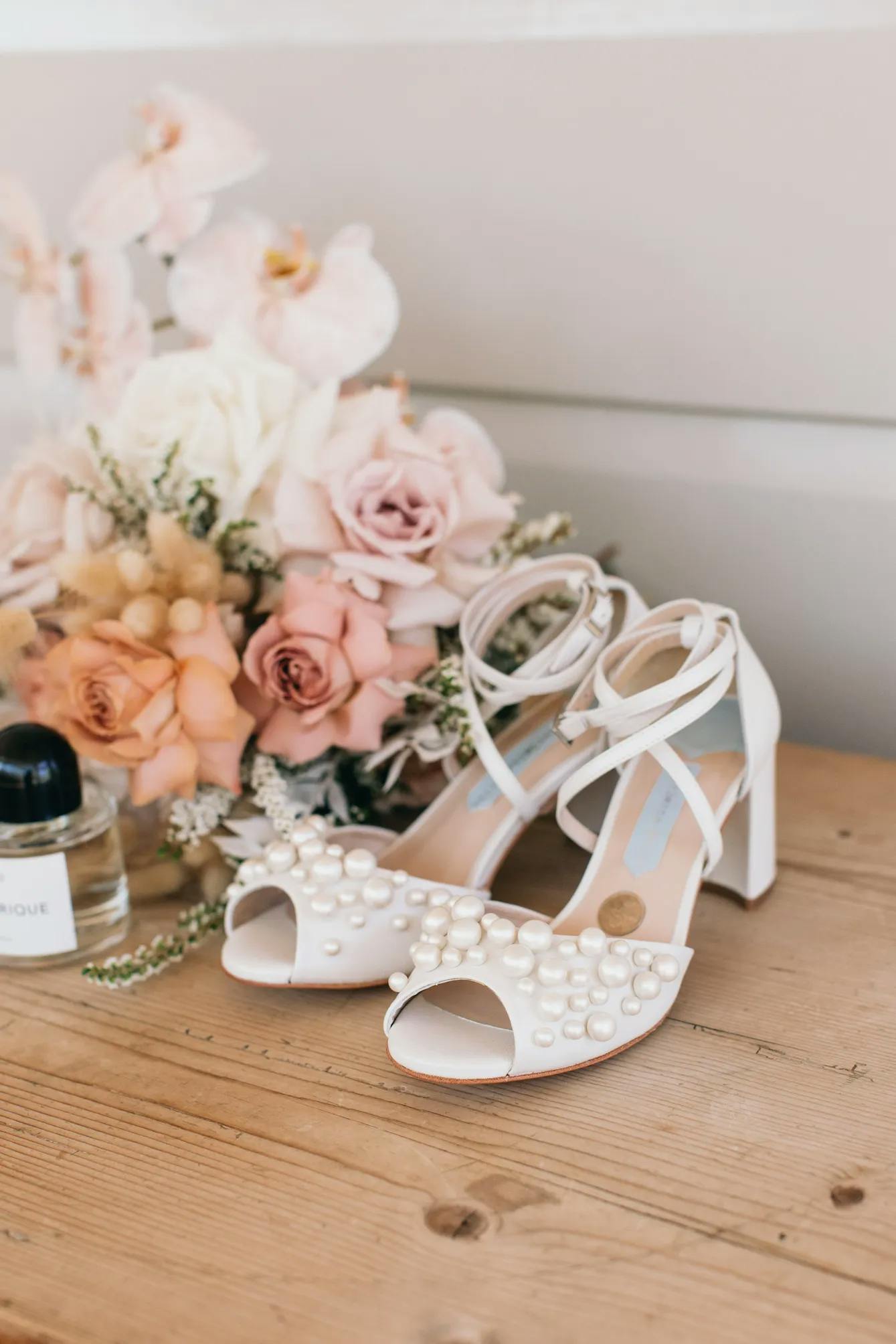 A pair of elegant white high-heeled sandals adorned with pearls is displayed on a wooden surface. Behind the shoes is a stylish floral arrangement with pink and white flowers, alongside a perfume bottle and a necklace, creating a delicate and chic composition.