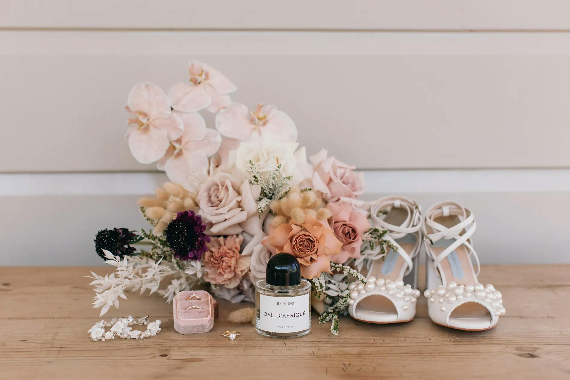 A wedding arrangement featuring pink and white flowers, a pair of white high-heeled shoes adorned with pearls, a bottle of perfume labeled "Bal d'Afrique," a pink velvet ring box with a ring inside, a pair of earrings, and a hairpin, all placed on a wooden surface.