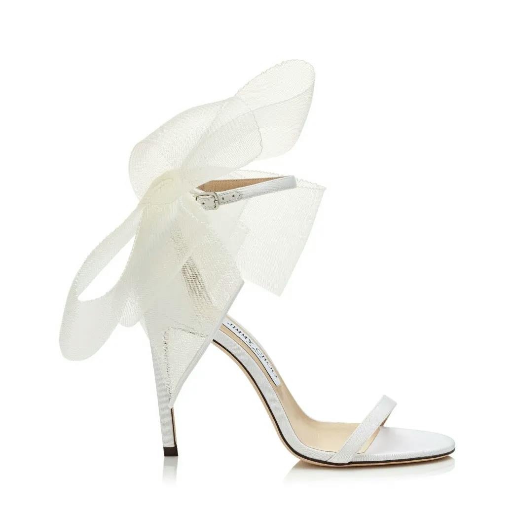 A white high-heeled sandal with a thin ankle strap, open toe, and a large, decorative, semi-transparent bow on the back. The sleek and elegant design is suitable for formal events or weddings.