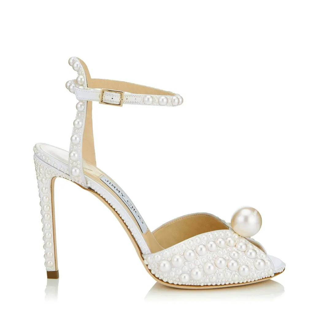 A high-heeled sandal decorated with white pearls. The shoe features a single strap around the ankle with a gold buckle, an open toe, and a pearl accent near the toe area. The shoe has a slender heel, also adorned with pearls. The interior is beige.