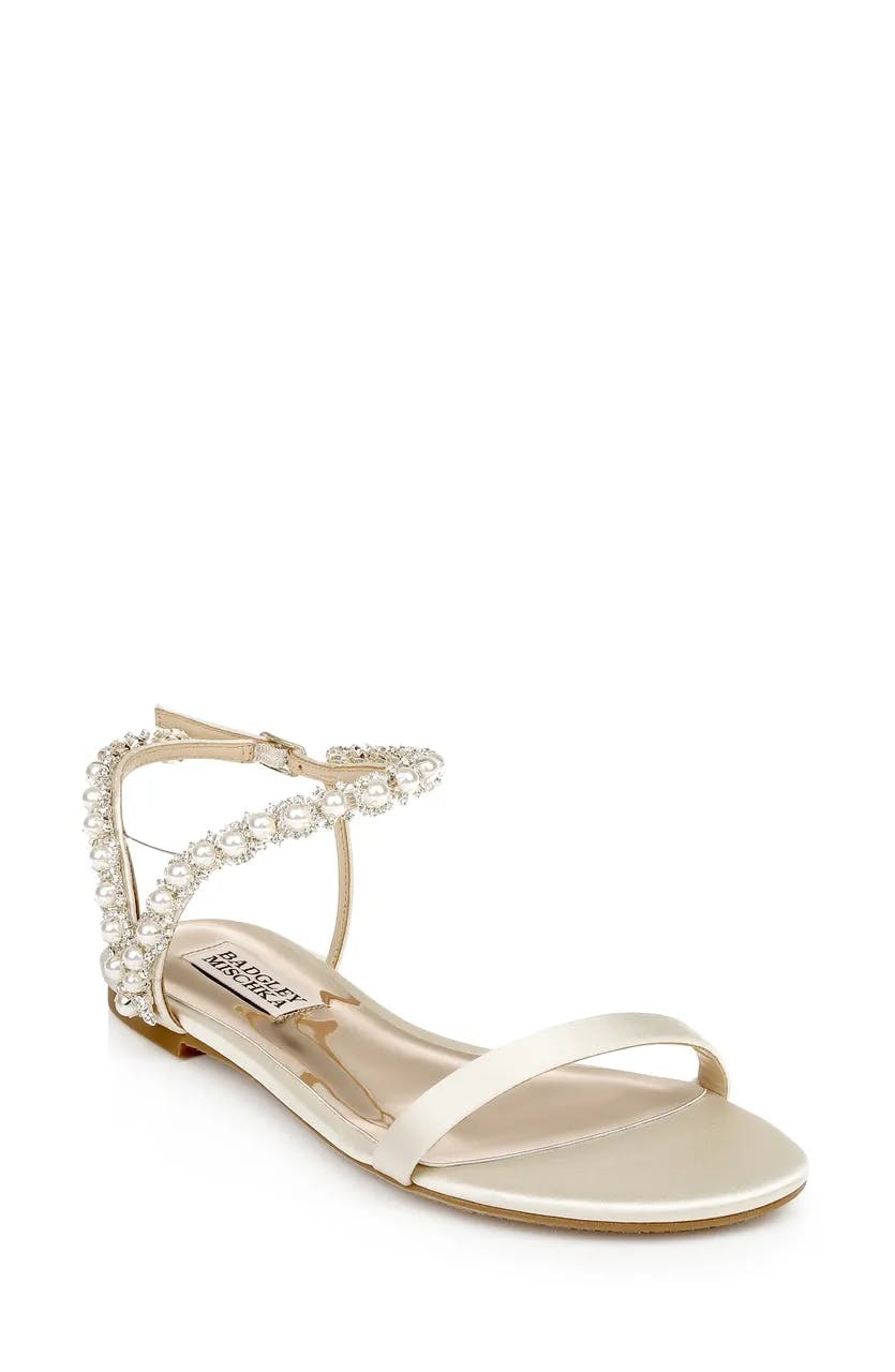 A single elegant sandal with a low heel, featuring a thin toe strap and an embellished ankle strap adorned with pearls and rhinestones. The sandal is beige in color and has a metallic accent on the sole.