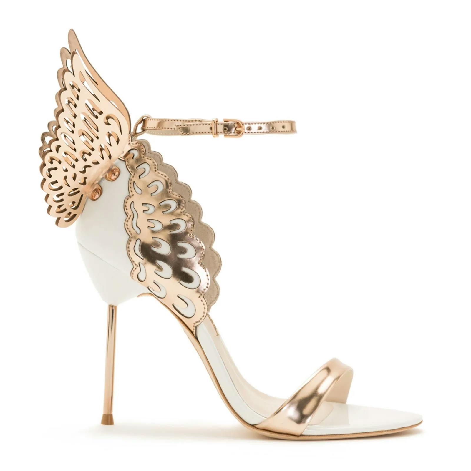 A metallic gold high-heeled sandal featuring intricate wing-like cutout designs on the heel and ankle strap. The elegant shoe has a thin stiletto heel, open toe, and a glossy finish, giving it a stylish and luxurious appearance.