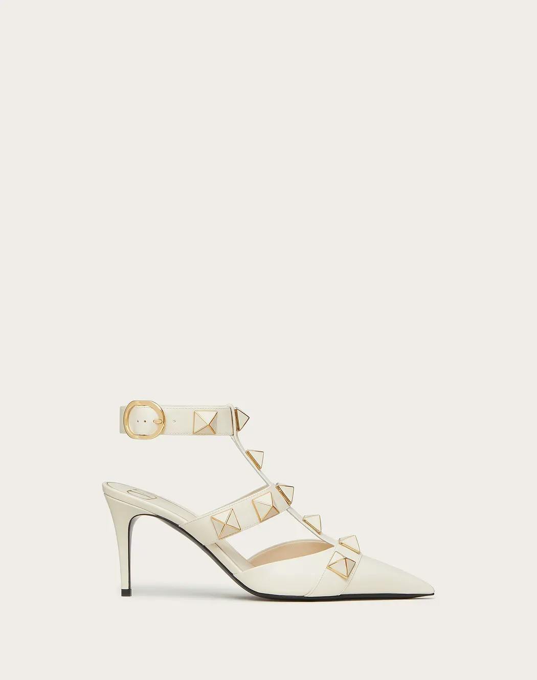 A stylish high-heeled shoe in white leather featuring a pointed toe design, pyramid stud embellishments, and an ankle strap with a buckle. The shoe has a mid-height stiletto heel and a T-strap connecting the toe area to the ankle strap.