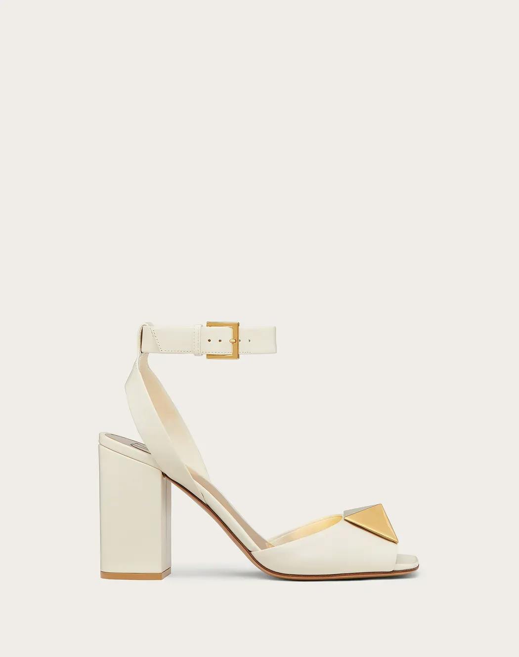 A white high-heeled sandal with a chunky heel, ankle strap with a gold buckle, and a gold triangular embellishment on the toe strap. The shoe has a minimalist and elegant design suitable for formal wear.