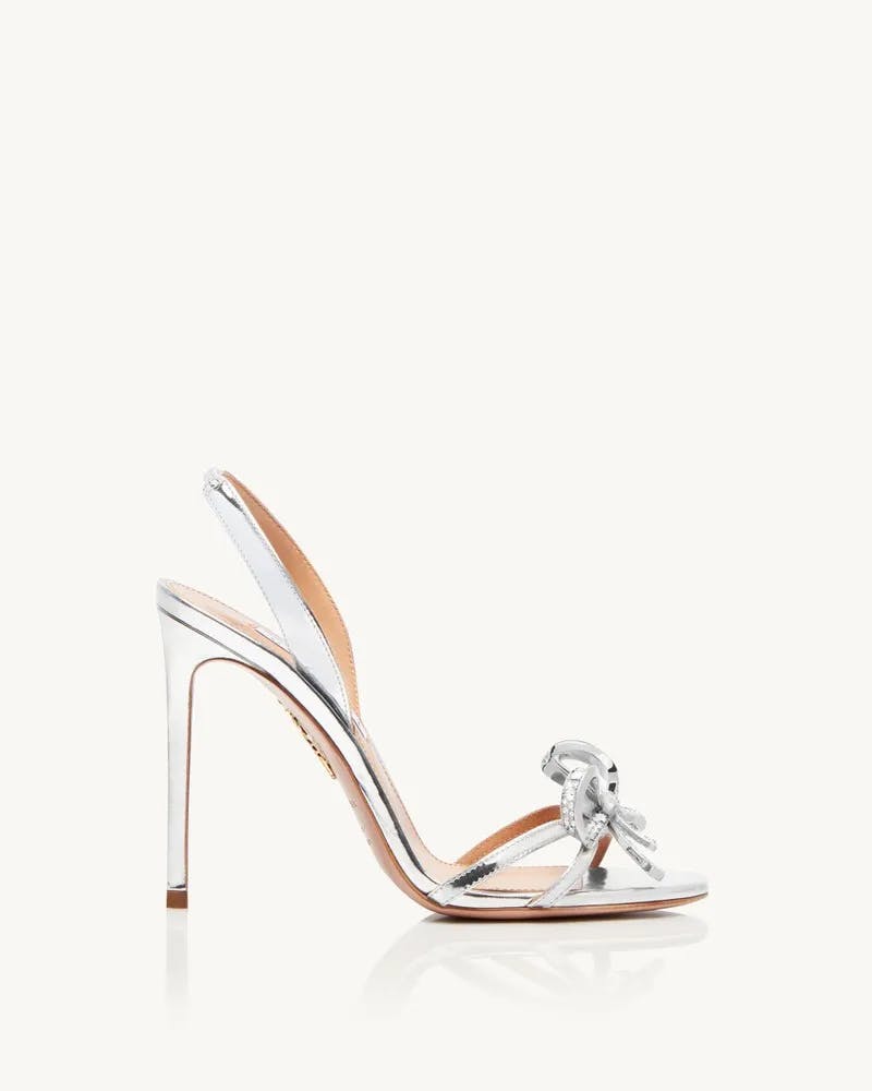 A high-heeled sandal with a shiny silver finish. The sandal features thin straps, one across the toes adorned with a large silver bow embellishment, and another around the heel. The shoe has a sleek, elegant design with a stiletto heel and a beige sole.
