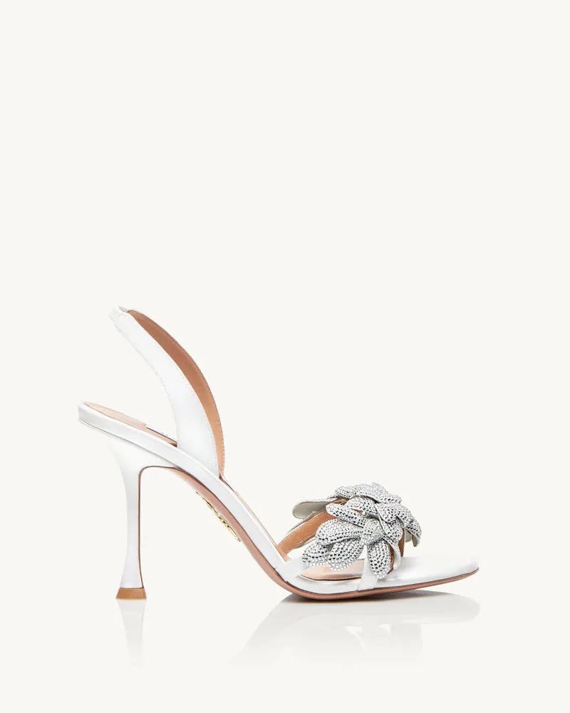 The image shows a white high-heeled sandal with an open toe design and an ankle strap. The sandal features an ornate silver flower embellishment on the toe strap. The heel is sleek and slender, adding an elegant touch to the sophisticated footwear.