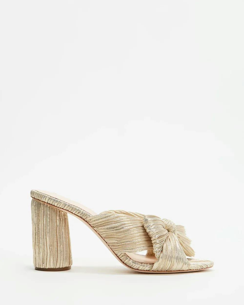 A single gold-toned high-heeled sandal is shown against a white background. The sandal has a thick, block heel and features a crinkled texture with a knot detail at the front strap. The heel and strap are made from the same textured material.
