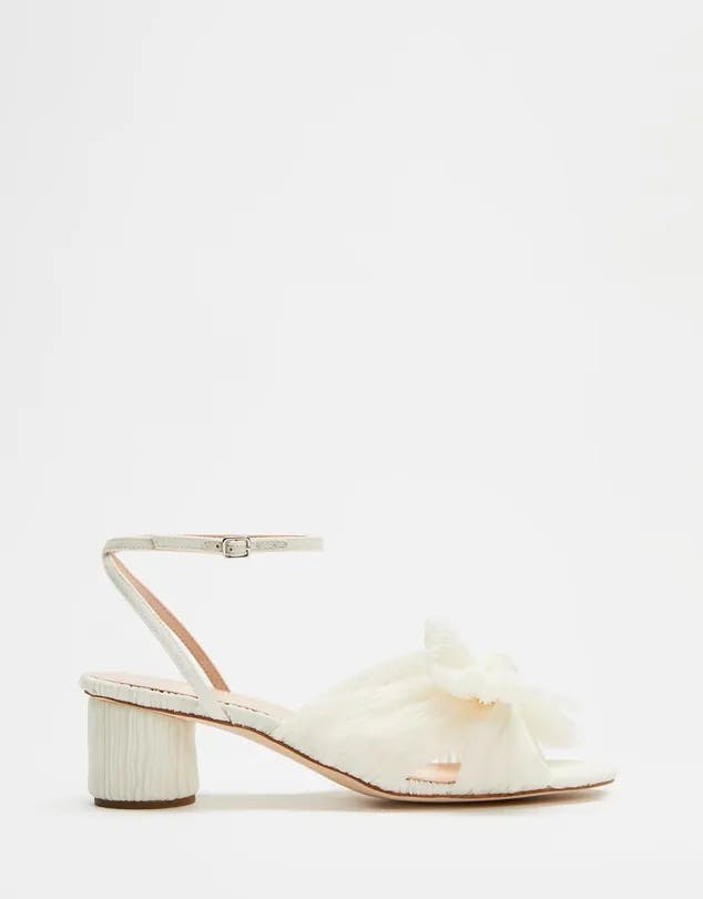 A white open-toe sandal with a block heel. It has a delicate ankle strap with a buckle and a large decorative bow on the front. The heel has a textured pattern while the insole and inner lining appear to be in a light pink shade.