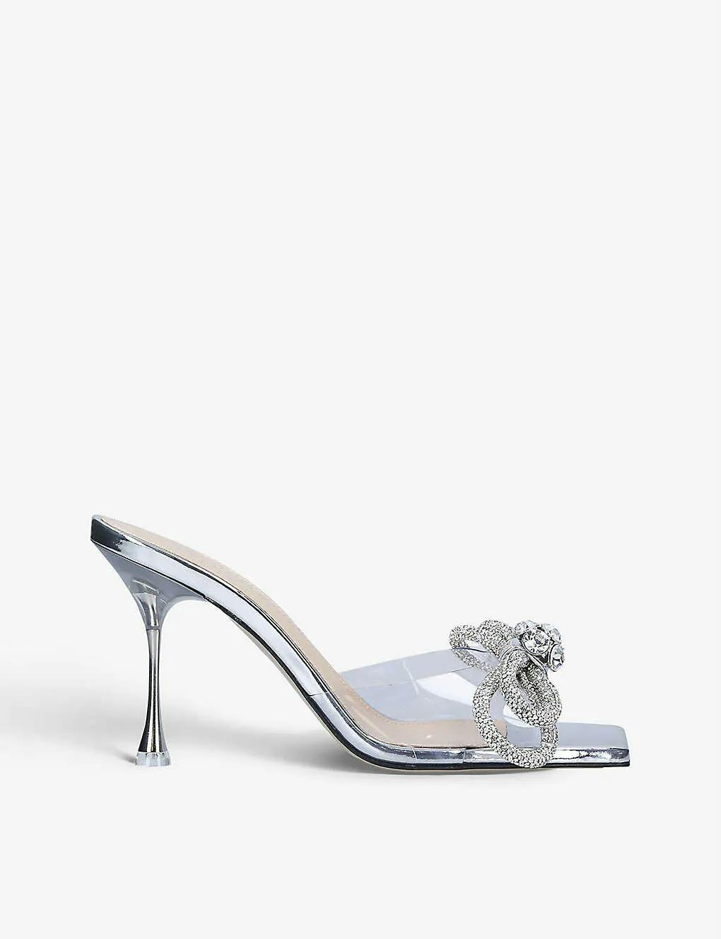 A high-heeled silver sandal with a clear strap adorned with a decorative, sparkling bow accent. The heel is thin and metallic, and the insole is a light beige color. The shoe is modern and elegant, suitable for dressy occasions.
