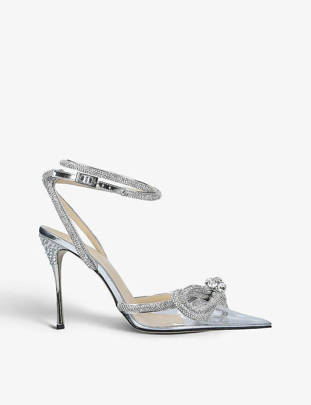 A silver high-heeled pump with a pointed toe, adorned with intricate rhinestones. The design features an ankle strap with sparkling embellishments and a decorative rhinestone bow on the front, creating an elegant and glamorous appearance.