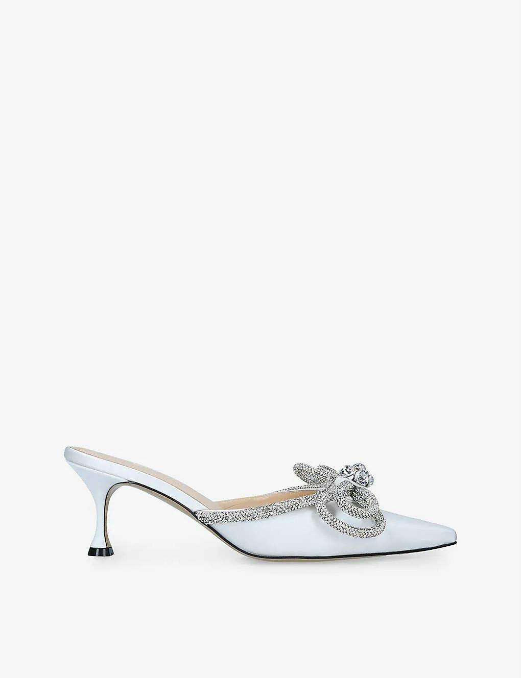 A stylish white satin mule with a pointed toe and a low, thin heel. The shoe features an elegant jeweled bow embellishment on the top, adorned with sparkling crystals. The background is plain white.