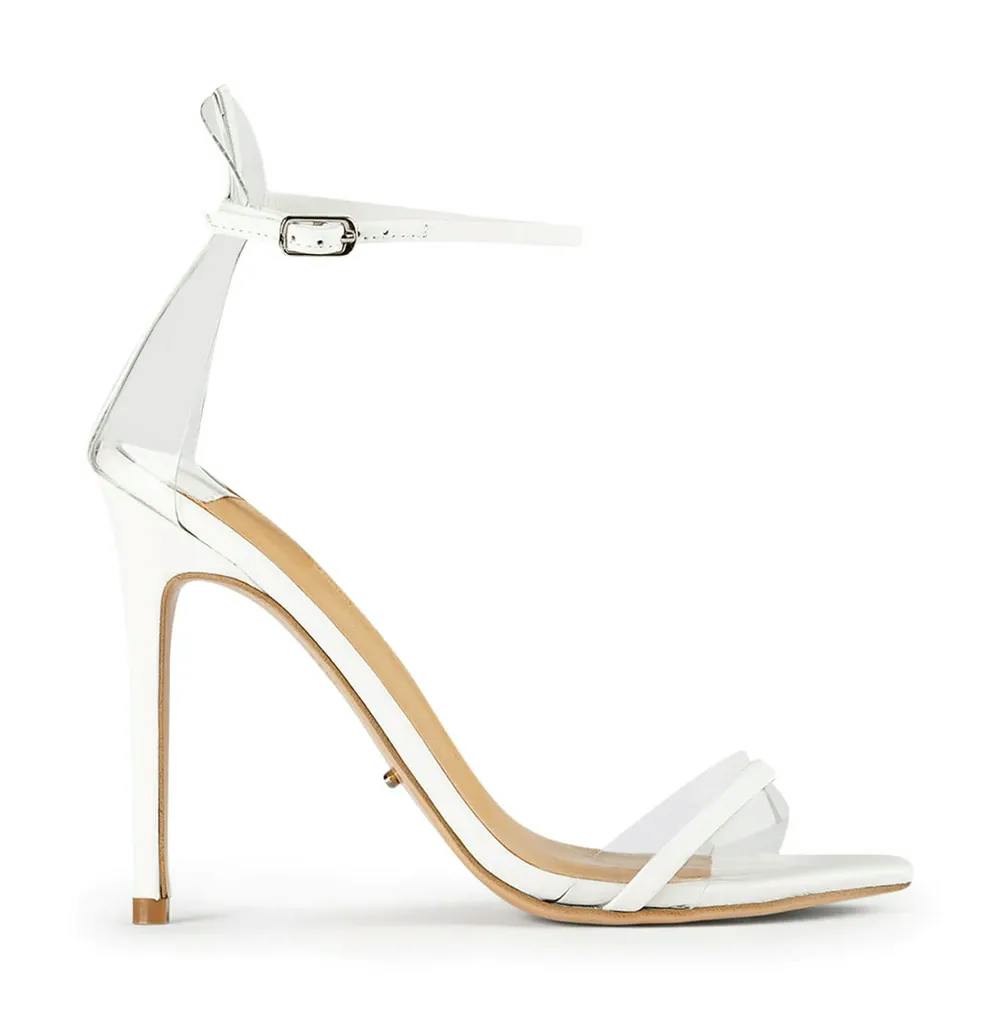 A white high-heeled sandal with a thin ankle strap and buckle closure. The shoe features a thin stiletto heel and a minimalist design with two slender straps across the toes. The interior sole appears beige. The sandal is set against a plain white background.