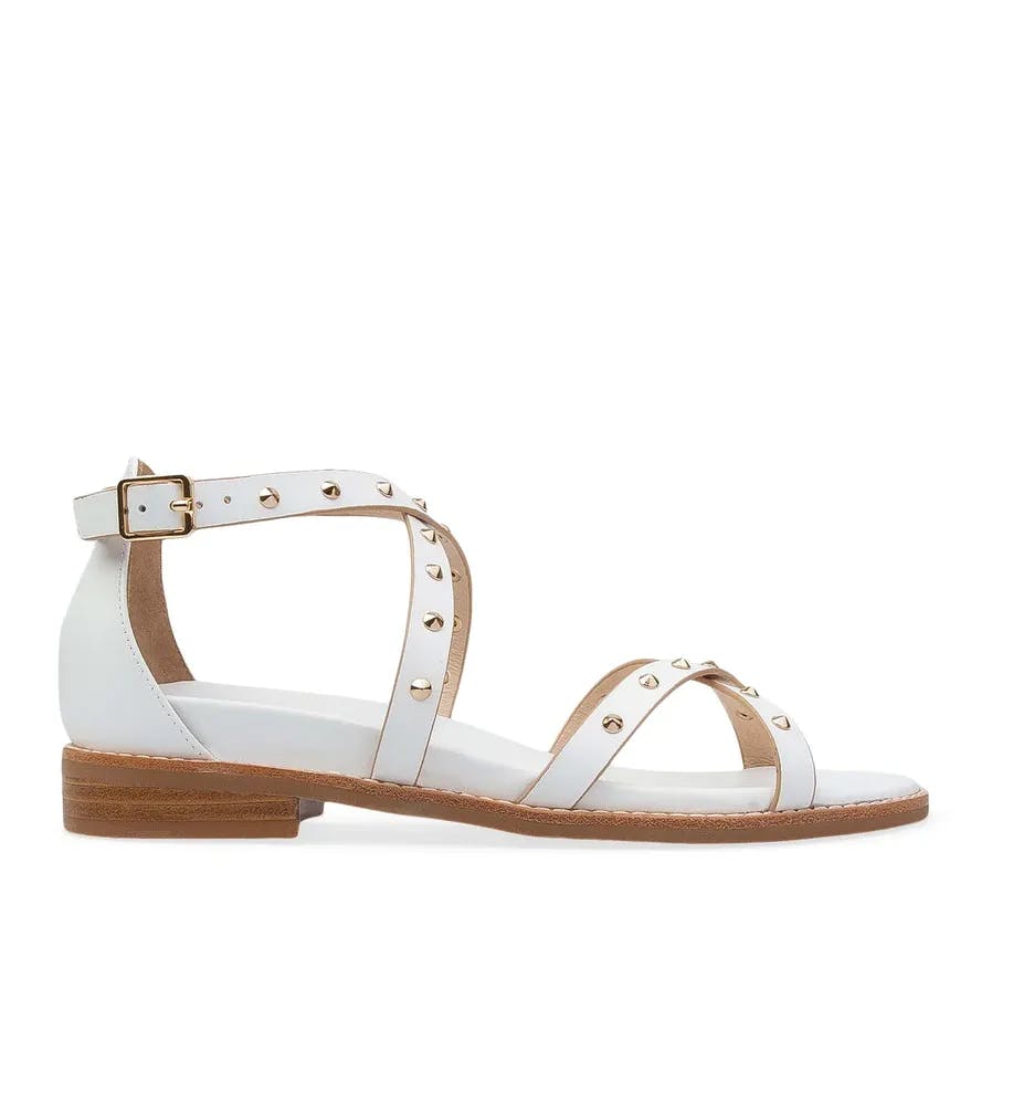 A white sandal with gold studs and a wooden low heel. The design includes an ankle strap with a buckle, a horizontal strap across the foot, and another strap across the toes. The sandal has a simple and elegant look suitable for casual or semi-formal wear.