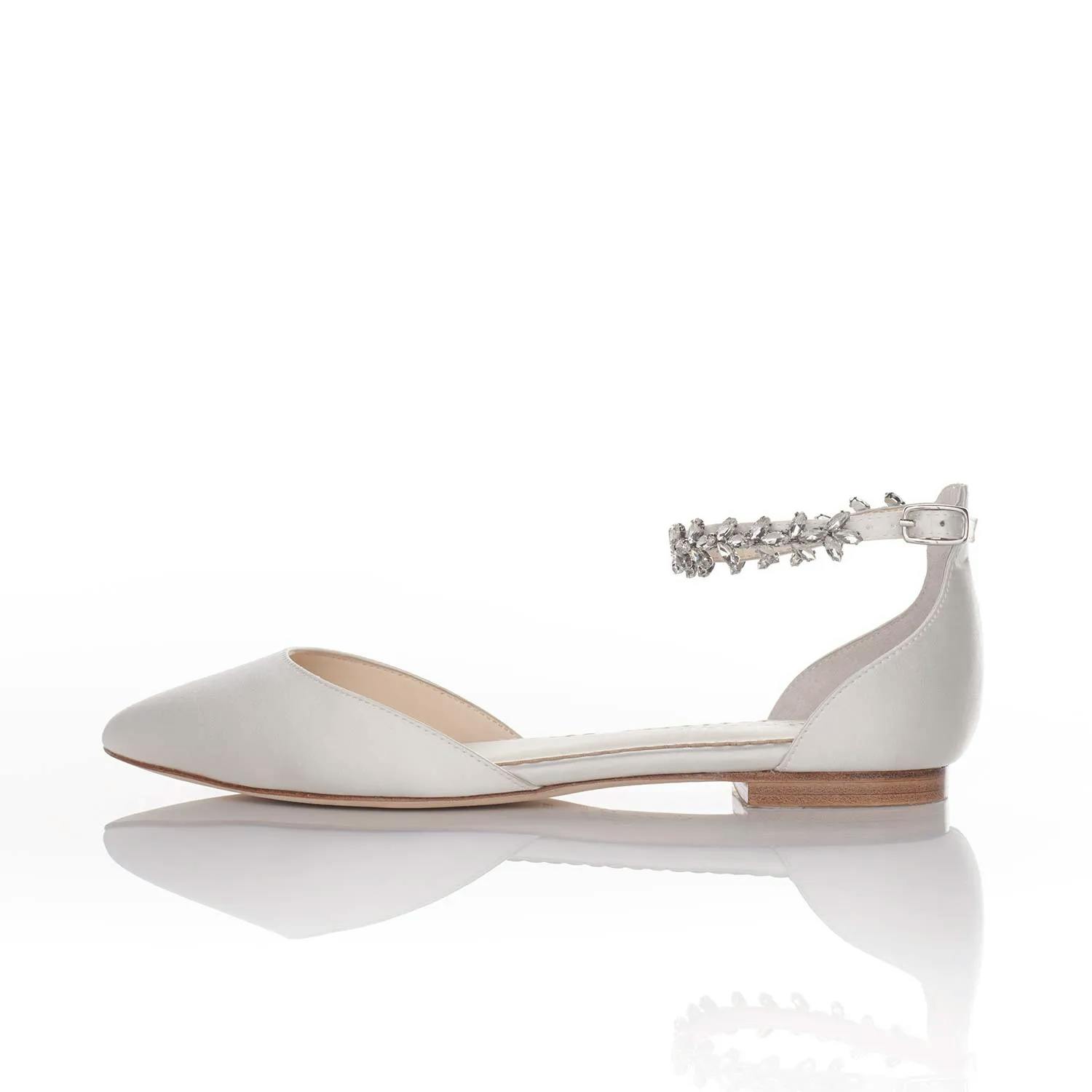 A white flat shoe with a closed pointed toe and a low wooden heel. The shoe features a decorative ankle strap adorned with intricate silver floral embellishments, fastened with a small buckle. The background is plain white.