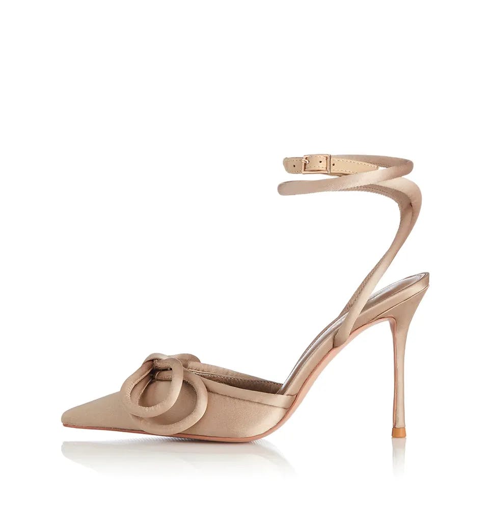 A stylish, beige stiletto heel shoe with a pointed toe. It features a decorative bow on the front and an elegant, thin ankle strap with a small buckle. The stiletto heel is high and slender, giving the shoe a sophisticated appearance.