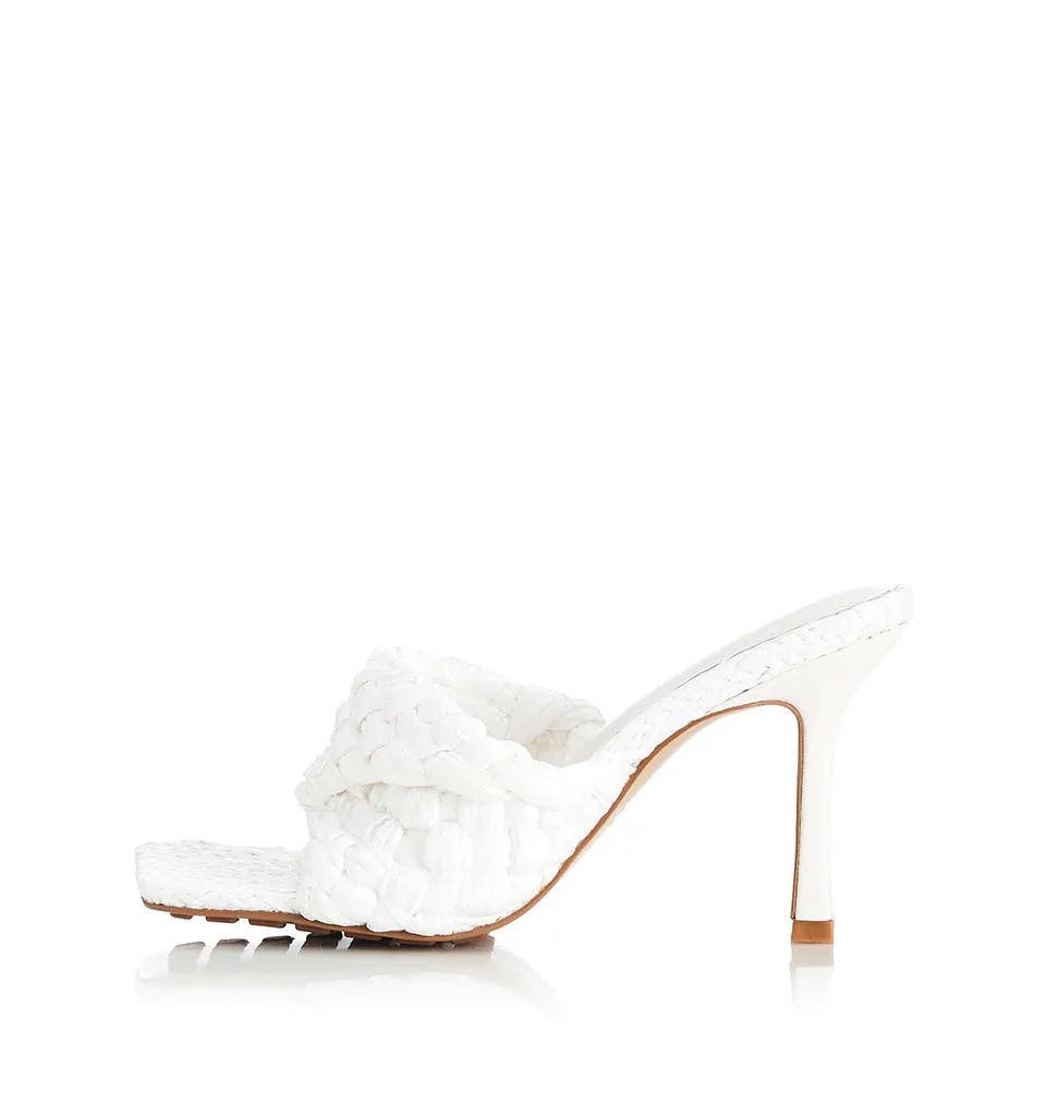 A single white high-heeled mule with a braided strap across the top and an open toe design. The heel is stiletto-style and the shoe features a textured pattern. The background is plain white.