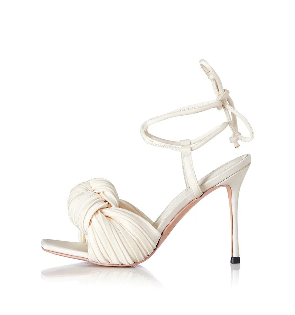 A stylish high-heeled sandal featuring a cream-colored, twisted knotted front strap design. The shoe has a delicate ankle strap that ties around the ankle and a sleek stiletto heel. The sole and inner lining are light pink.