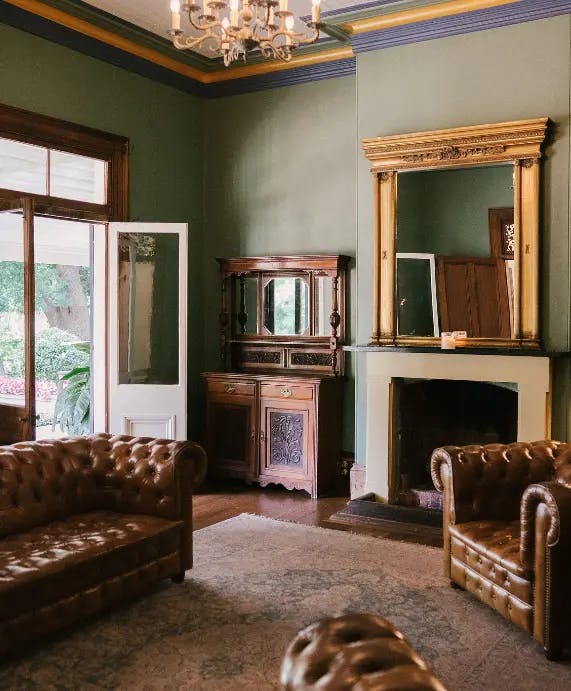 An elegant room features green walls, a large mirror above a fireplace, a wooden sideboard with a mirror, and two brown leather chesterfield sofas. A chandelier hangs from the ceiling, while a white door opens to a view of greenery outside. An area rug covers part of the wooden floor.
