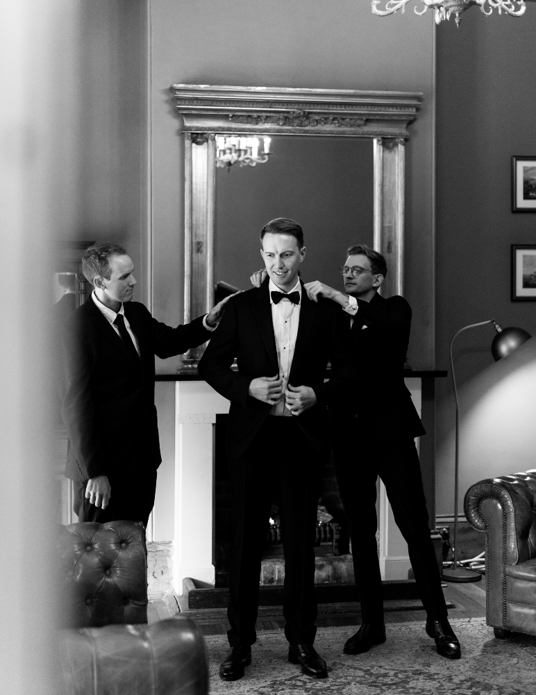 A black-and-white photo of a groom in a tuxedo, standing in front of a fireplace with a large mirror above it. Two groomsmen, also in tuxedos, are adjusting his jacket and bow tie, preparing for the wedding ceremony. The setting is an elegant, classic room.