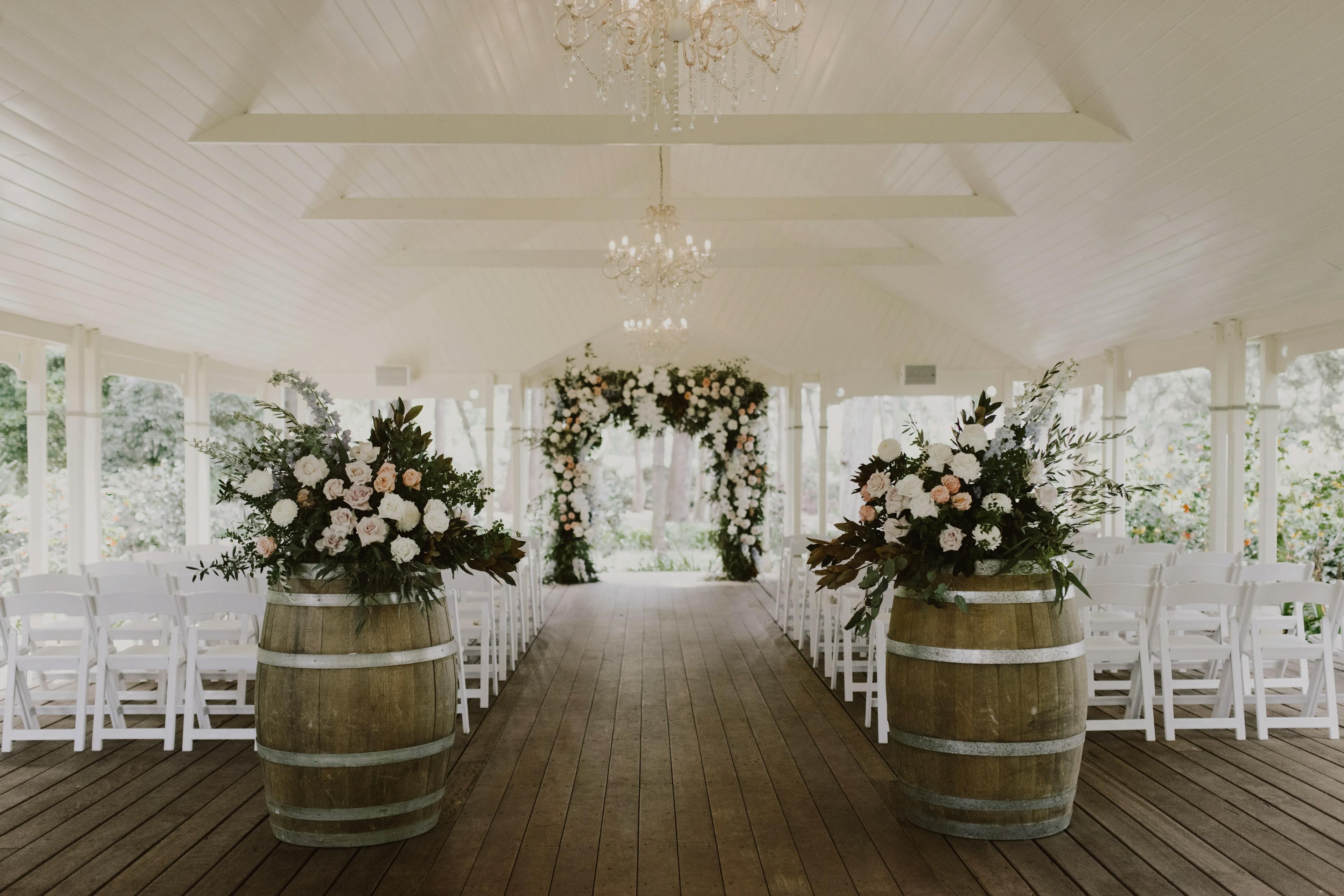 A beautifully decorated indoor wedding venue with wooden flooring and white ceilings. Two large floral arrangements sit on top of wooden barrels at the aisle's entrance, leading to a white arch adorned with flowers. White chairs are arranged on both sides of the aisle.