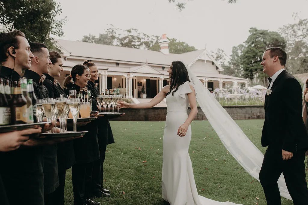 A bride and groom, both elegantly dressed, stand on a lawn in front of a large house. The bride, in a white dress with a long veil, takes a drink from a tray held by a server. Several servers dressed in black uniforms hold trays with beverages, lined up in a row.