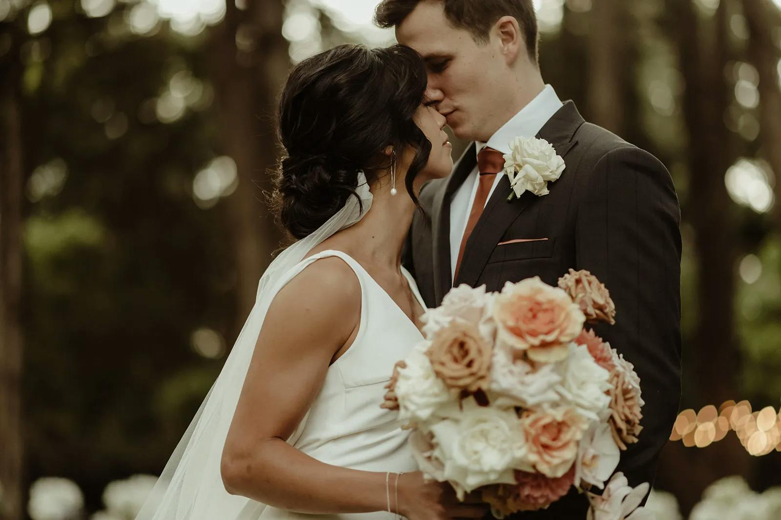 A bride and groom embrace tenderly in a wooded outdoor setting. The bride wears a white dress and holds a large bouquet of pink, peach, and white flowers. The groom, in a dark suit with a white rose boutonniere, gently kisses the bride on the forehead.