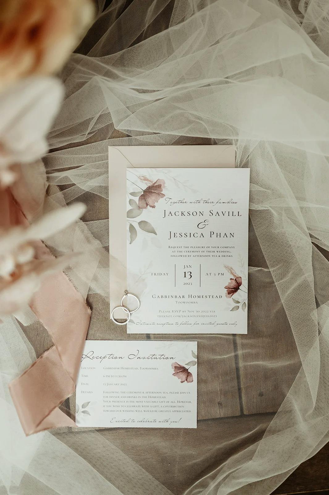 A wedding invitation set on a draped, sheer fabric. The invitation features names, "Jackson Savill & Jessica Phan," and details for the event on January 13th at Gardiner Homestead. Also shown are wedding rings and a smaller reception card.