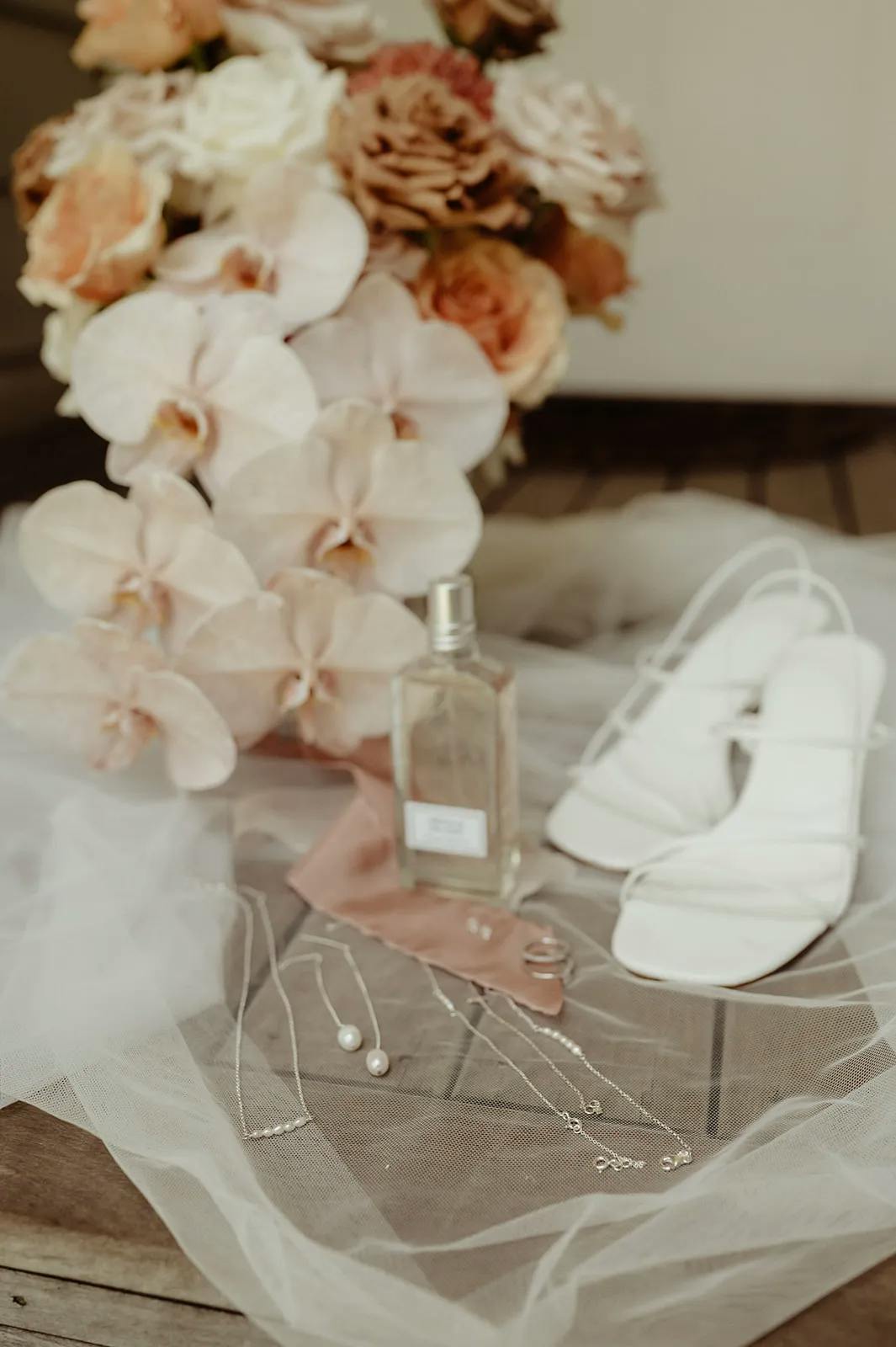 A wedding scene featuring a bouquet of flowers with cream and peach roses and orchids, a pair of white strappy high heels, a bottle of perfume, and various pieces of delicate jewelry including necklaces, earrings, and rings, all arranged on a soft veil.