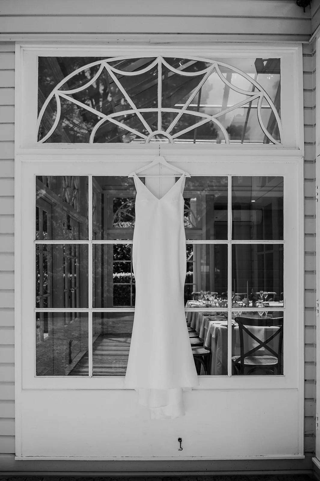 A white sleeveless wedding dress hangs elegantly on a large window with decorative trim. The window reveals a beautifully set dining area inside, with neatly arranged tables and chairs. The scene is captured in black and white.