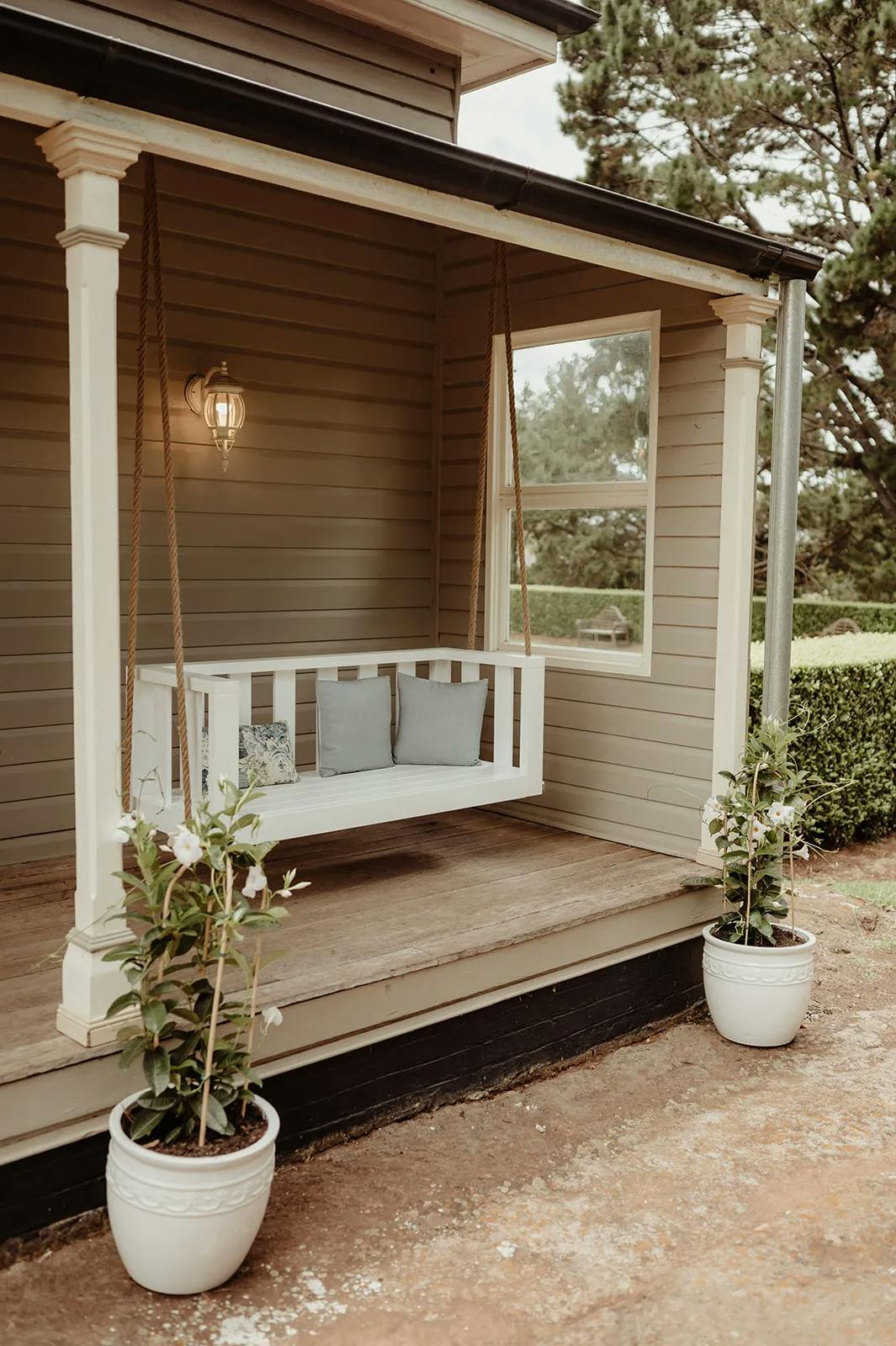 A cozy porch with a hanging white wooden bench swing adorned with gray cushions. Two potted plants with white flowers flank the entrance, and a lantern-style light fixture is mounted on the wall. The background features trees and greenery.