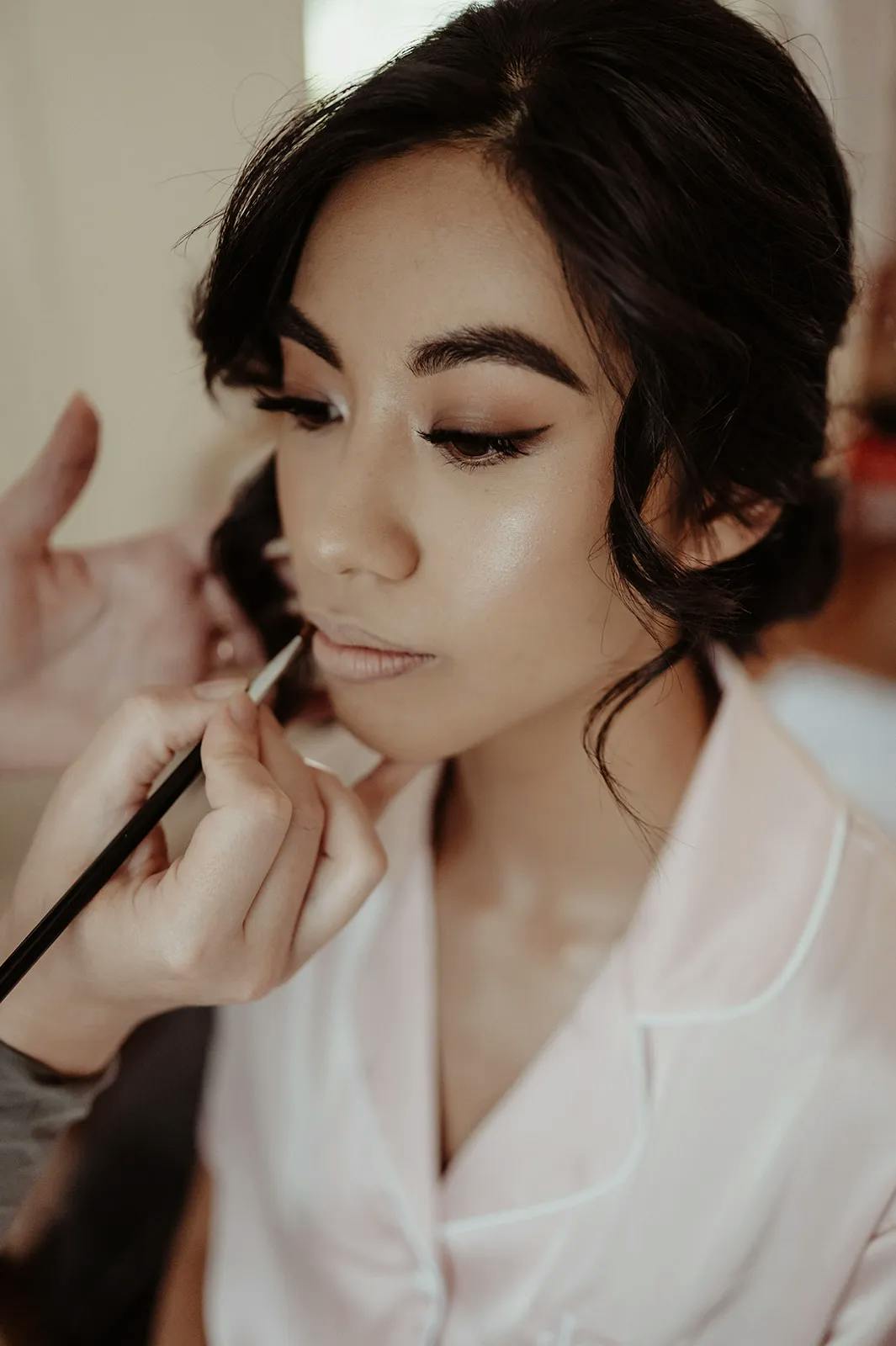 A woman with dark hair is getting her makeup done by another person. She is wearing a light pink top and her hair is styled in loose curls. The makeup artist is applying lipstick with a brush. The woman's eyes are closed and she appears calm.