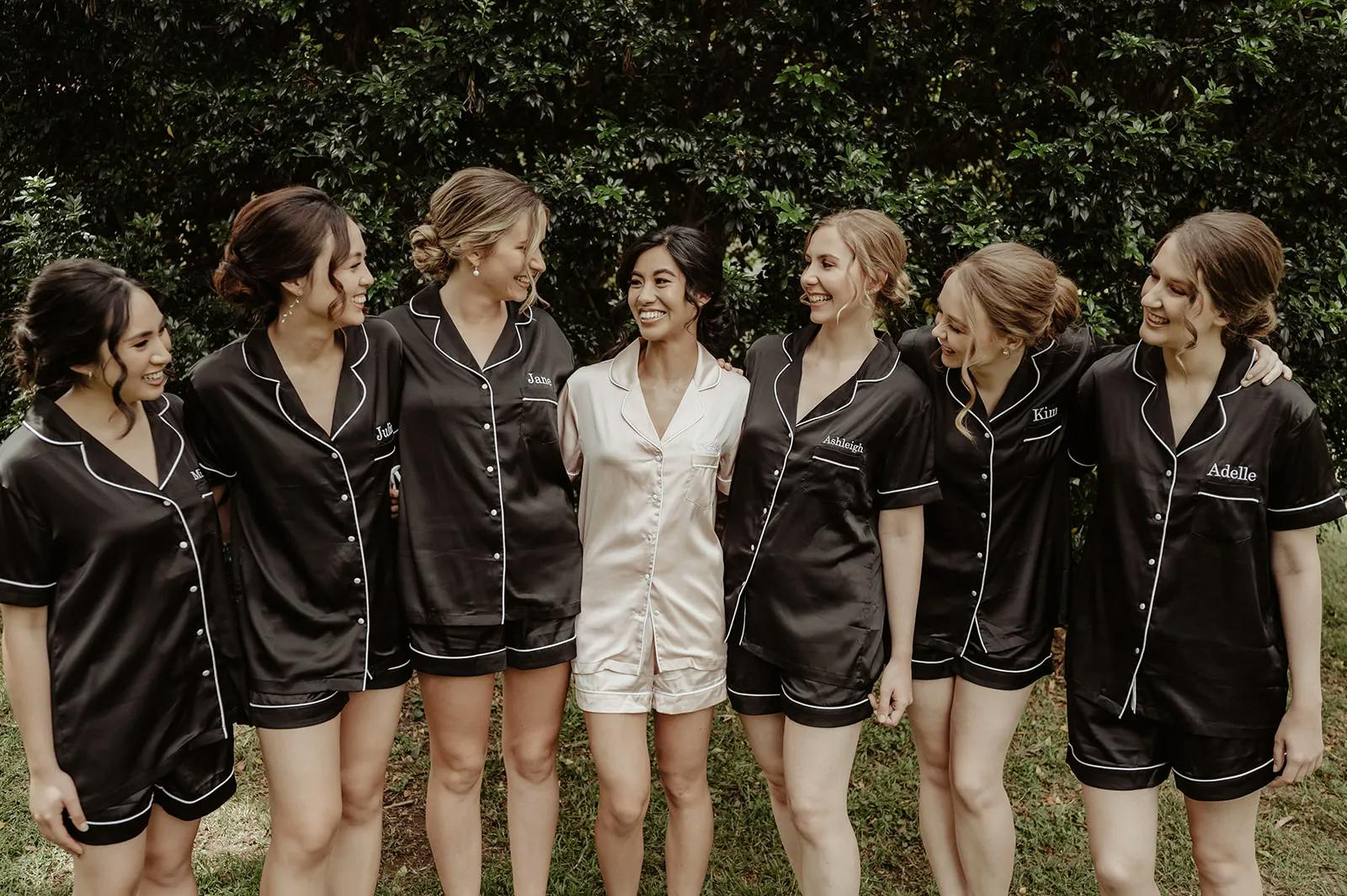 Seven women stand together outdoors, smiling and wearing matching pajamas. Six women wear black pajamas while one, in the center, wears white. They stand with arms around each other in front of a lush, green background.