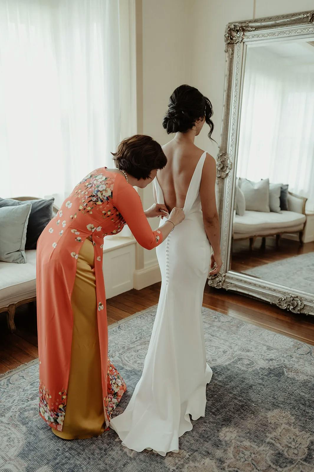 A woman in a white wedding dress stands in front of a full-length mirror while another woman, dressed in a floral-patterned reddish-orange and yellow gown, helps button up the back of the dress. The room has a cozy ambiance with sofas and soft natural light.