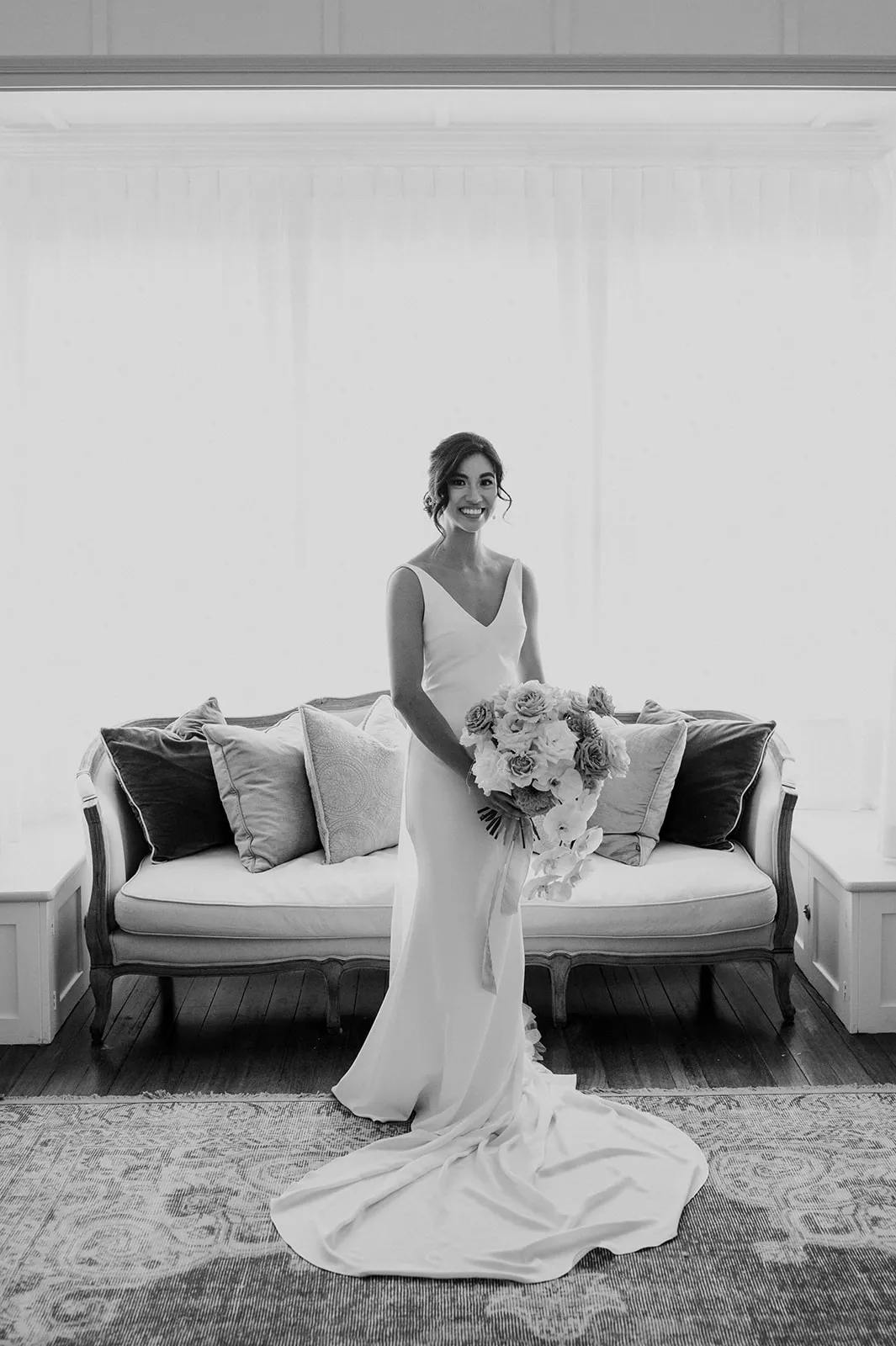 A bride in a flowing wedding gown stands in front of a sofa with multiple pillows. She holds a large bouquet of flowers, and the room is elegantly decorated with curtains and a patterned rug. The black and white photo captures her poised and smiling.
