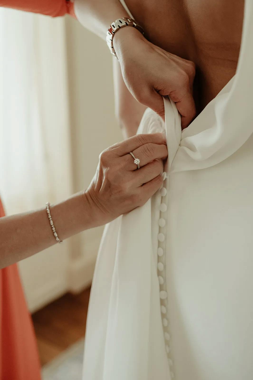 A person is helping another person by fastening buttons on the back of their white dress. The assisting person has a bracelet and a ring on their left hand. Only the hands and part of the torso of the person being helped are visible.