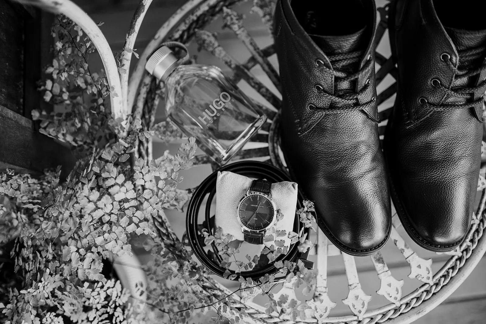 Black and white image of leather boots, a wristwatch on a cushion, a perfume bottle labeled "Hugo," and green foliage arranged on a perforated metal chair.