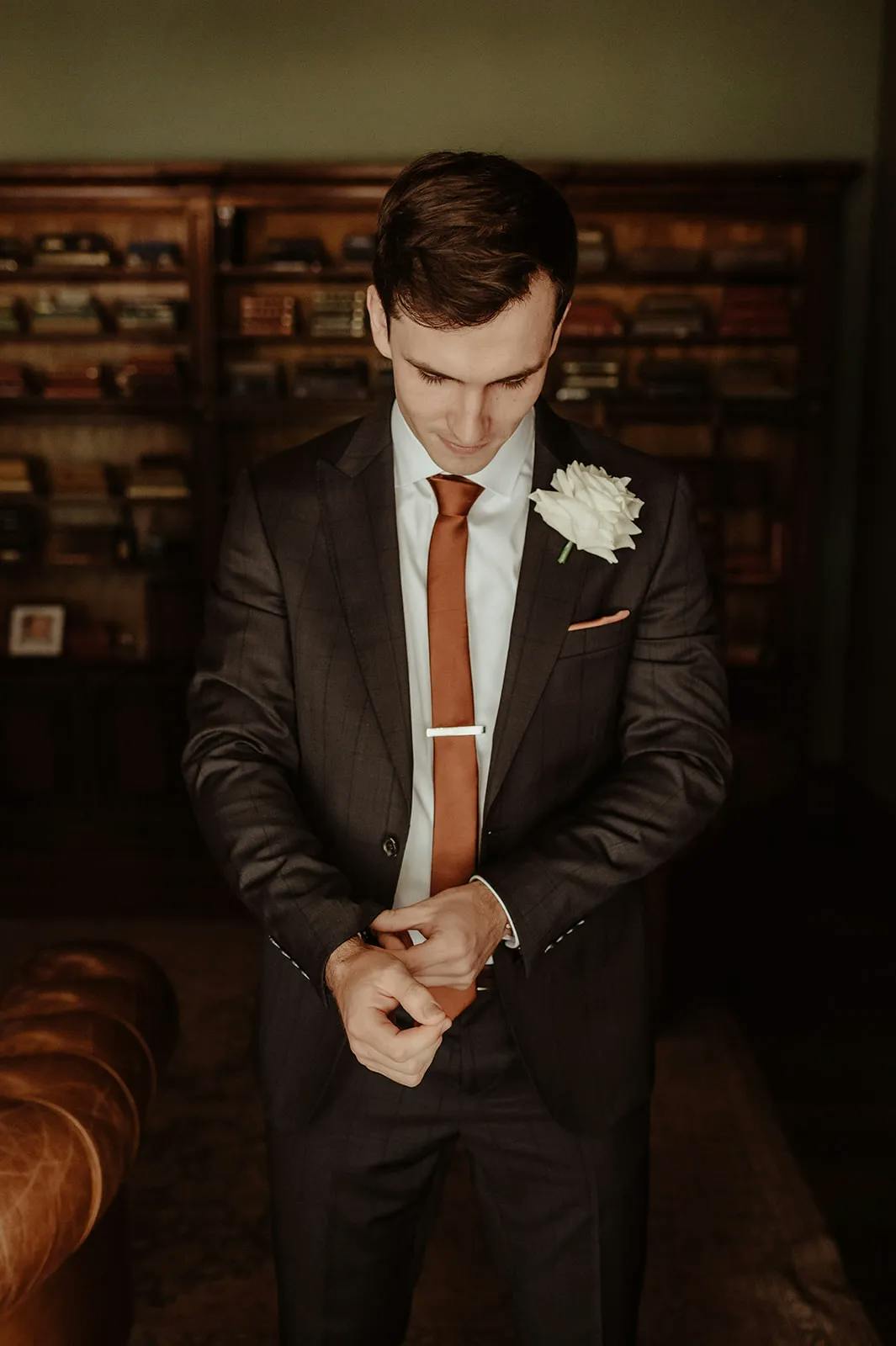 A man in a dark suit with a white boutonniere stands indoors, adjusting his cufflinks. He has a copper-colored tie and a tie clip. Behind him is a wooden bookshelf filled with books. The lighting is warm, creating a sophisticated and elegant atmosphere.