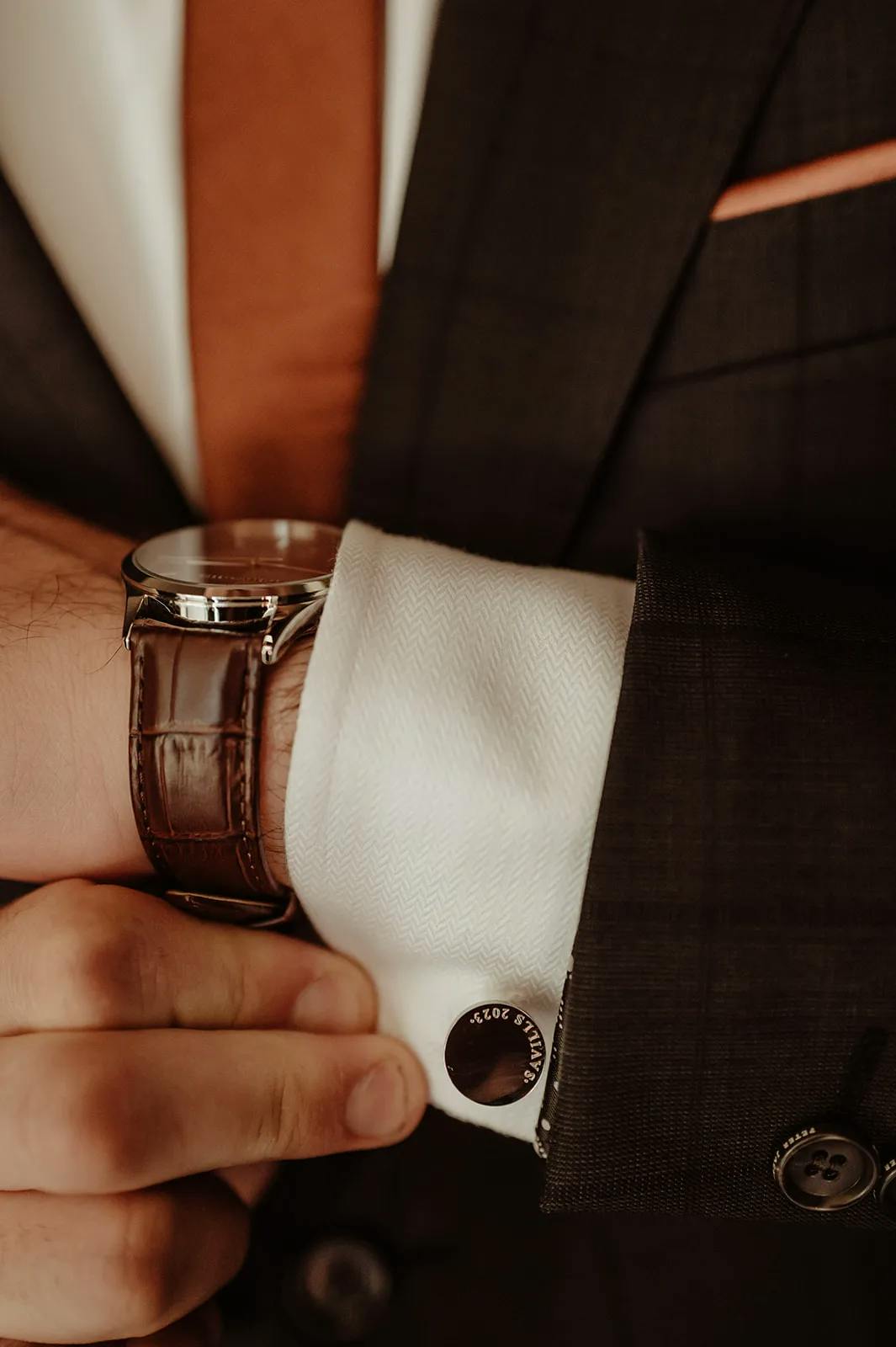 A close-up shot of a person in a dark suit with a white shirt and brown tie, adjusting their cufflinks. They are also wearing a watch with a brown leather strap and a visible silver dial.