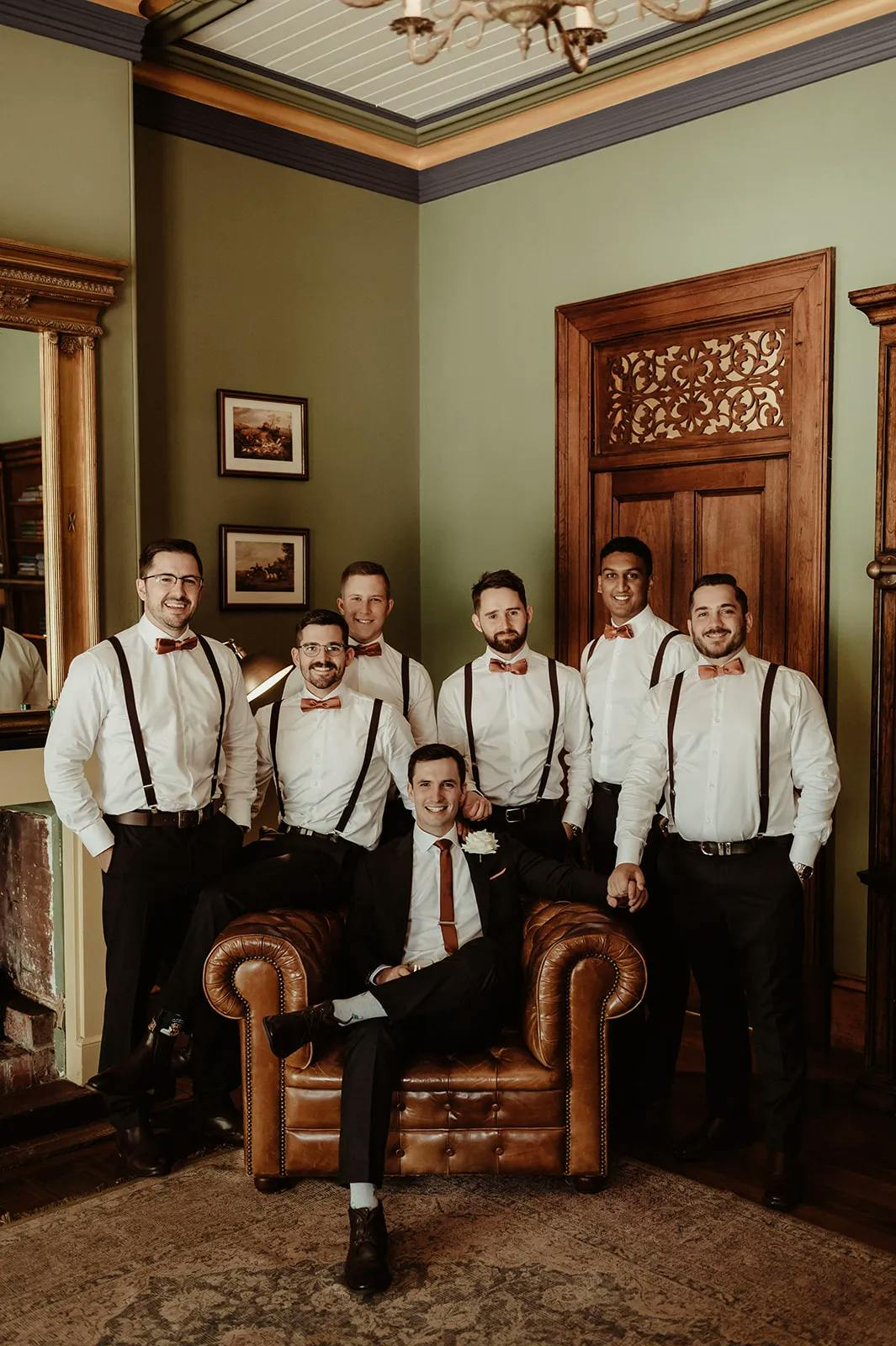 A group of seven men pose in a vintage-styled room. Six men stand behind a seated man, who is dressed in a suit with a red tie, while the standing men wear white shirts with suspenders. The room has green walls, wooden accents, and framed pictures on the wall.