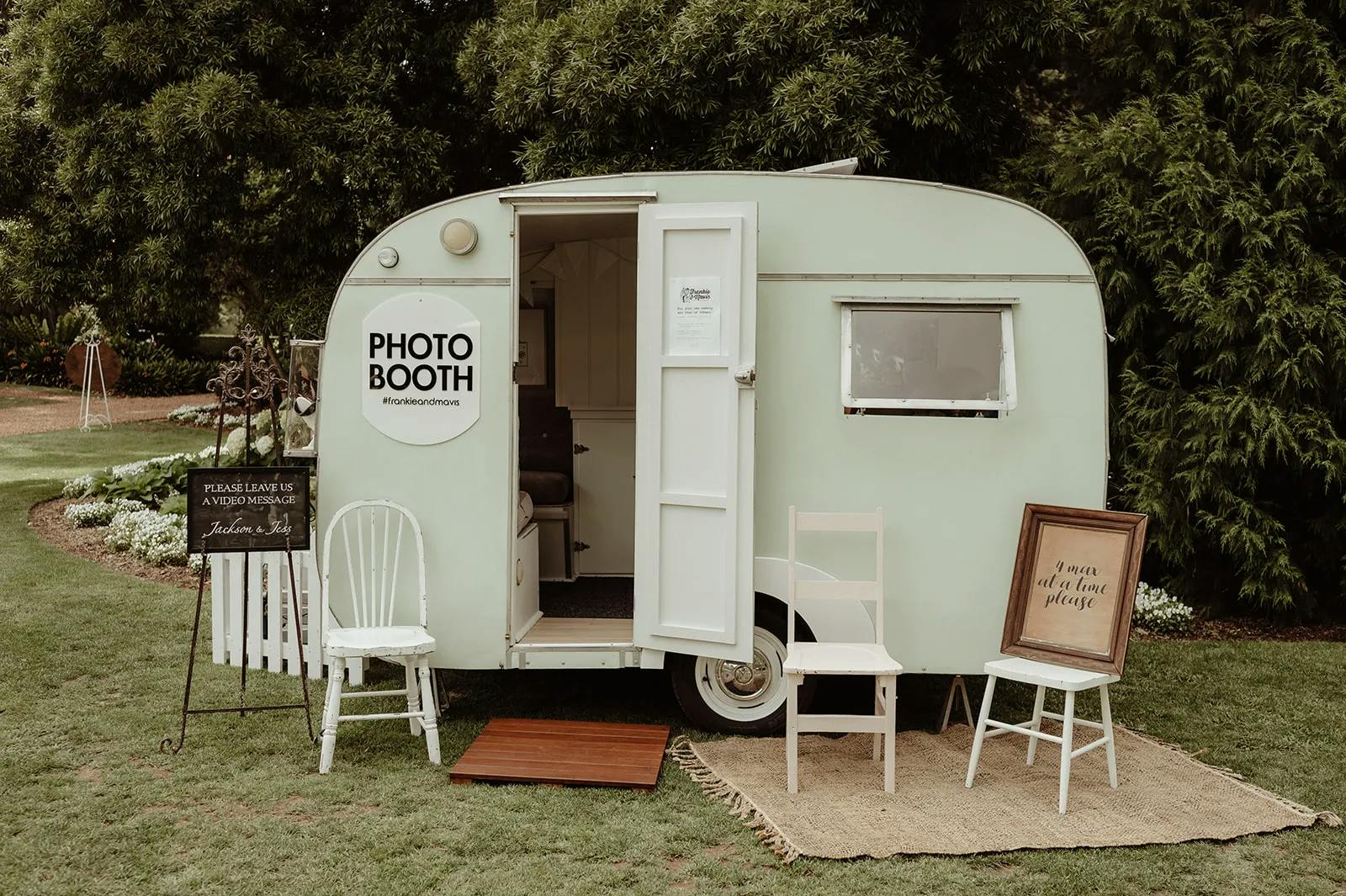 A pastel-colored trailer set up as a photo booth stands on a grassy area with trees in the background. In front, there are three chairs and a sign reading "In case you need a seat, please" on a rug. A signboard and a small table with props are placed nearby.