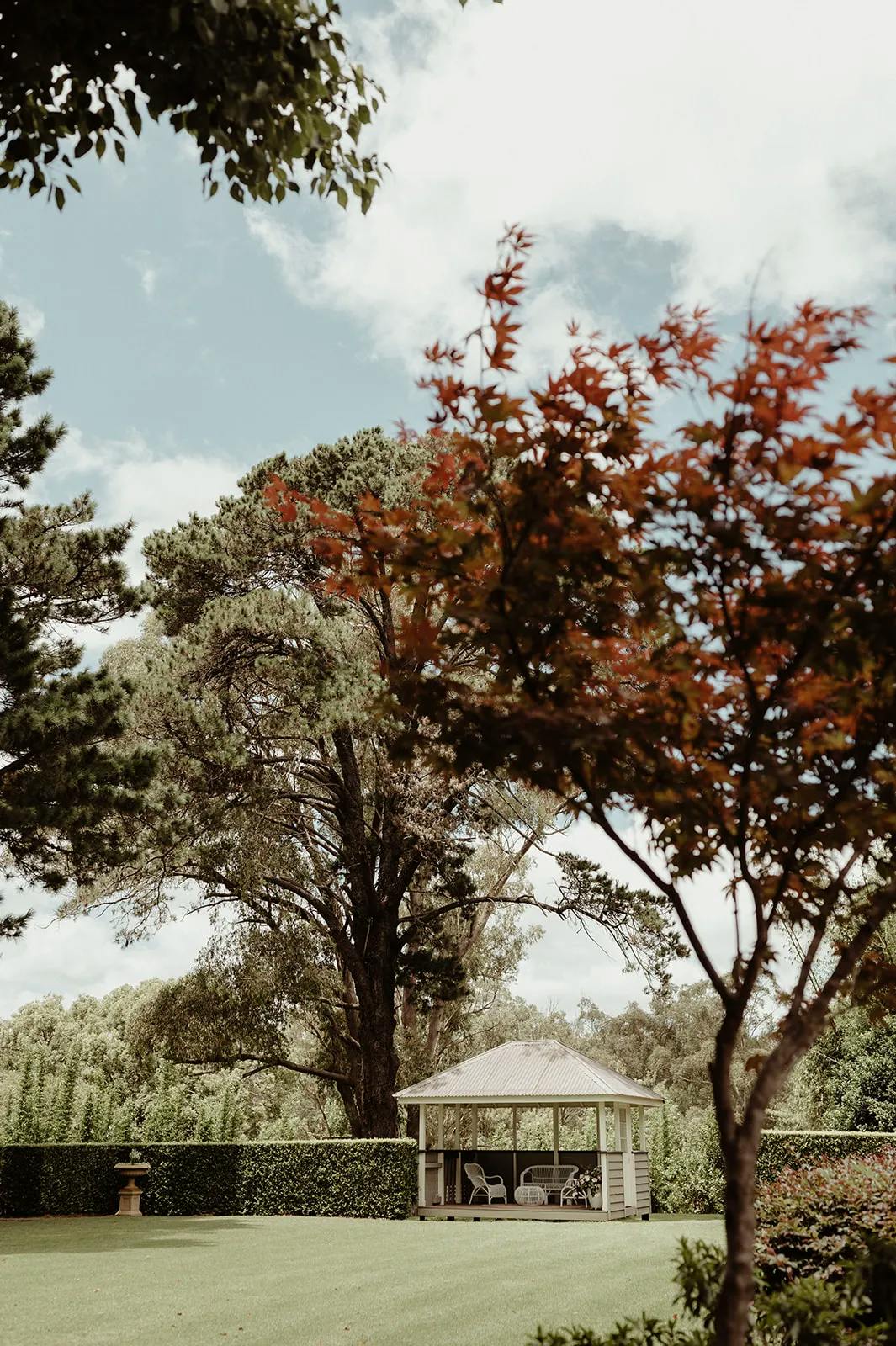 A serene garden scene featuring a white gazebo surrounded by lush greenery and tall trees. In the foreground, a tree with red leaves adds a splash of color. The sky is partly cloudy, casting dappled light on the peaceful landscape.